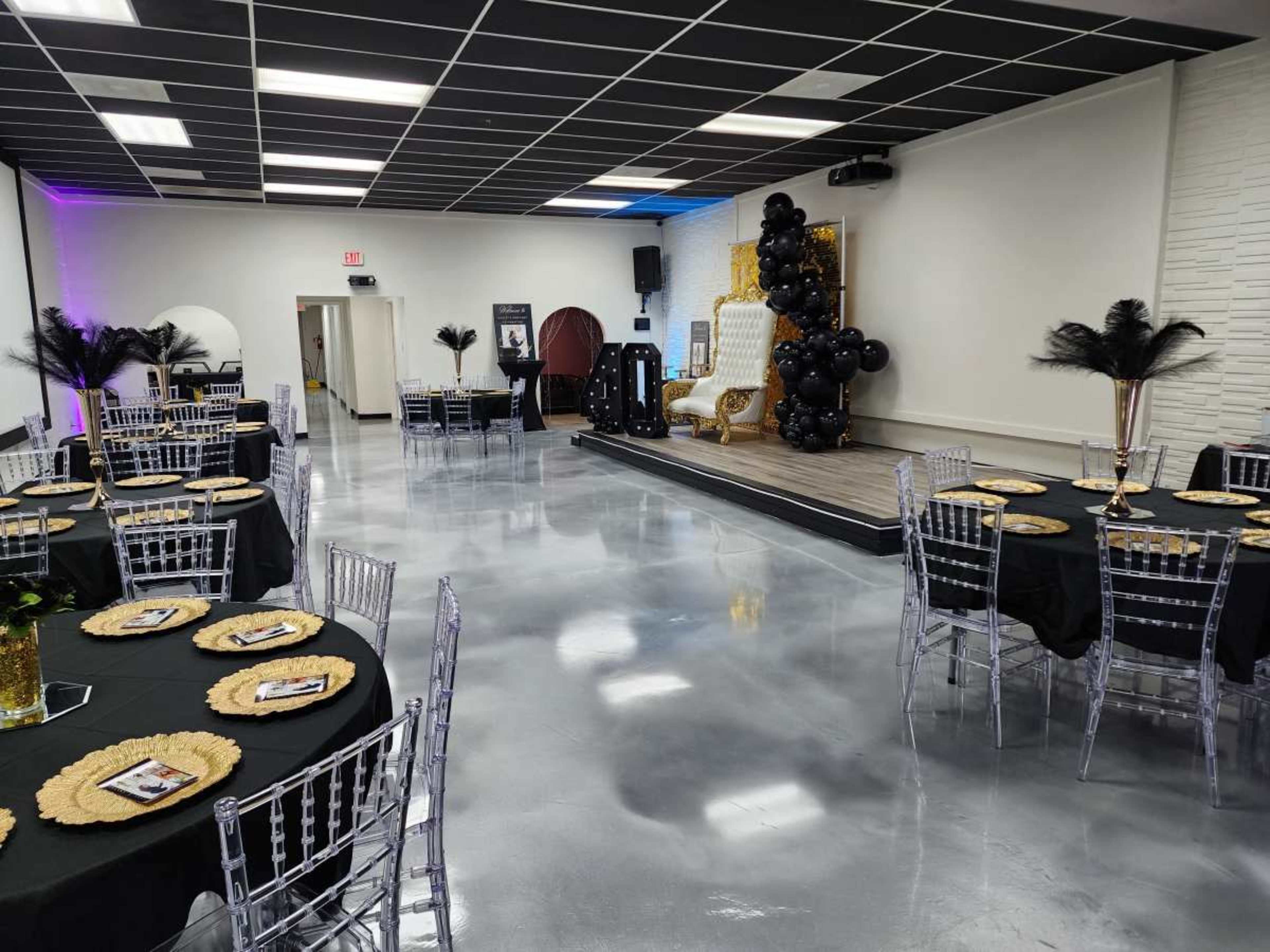 The image shows an elegantly arranged event space with round tables covered in black tablecloths and clear acrylic chairs, accented by a small stage featuring a decorated backdrop and black balloons.