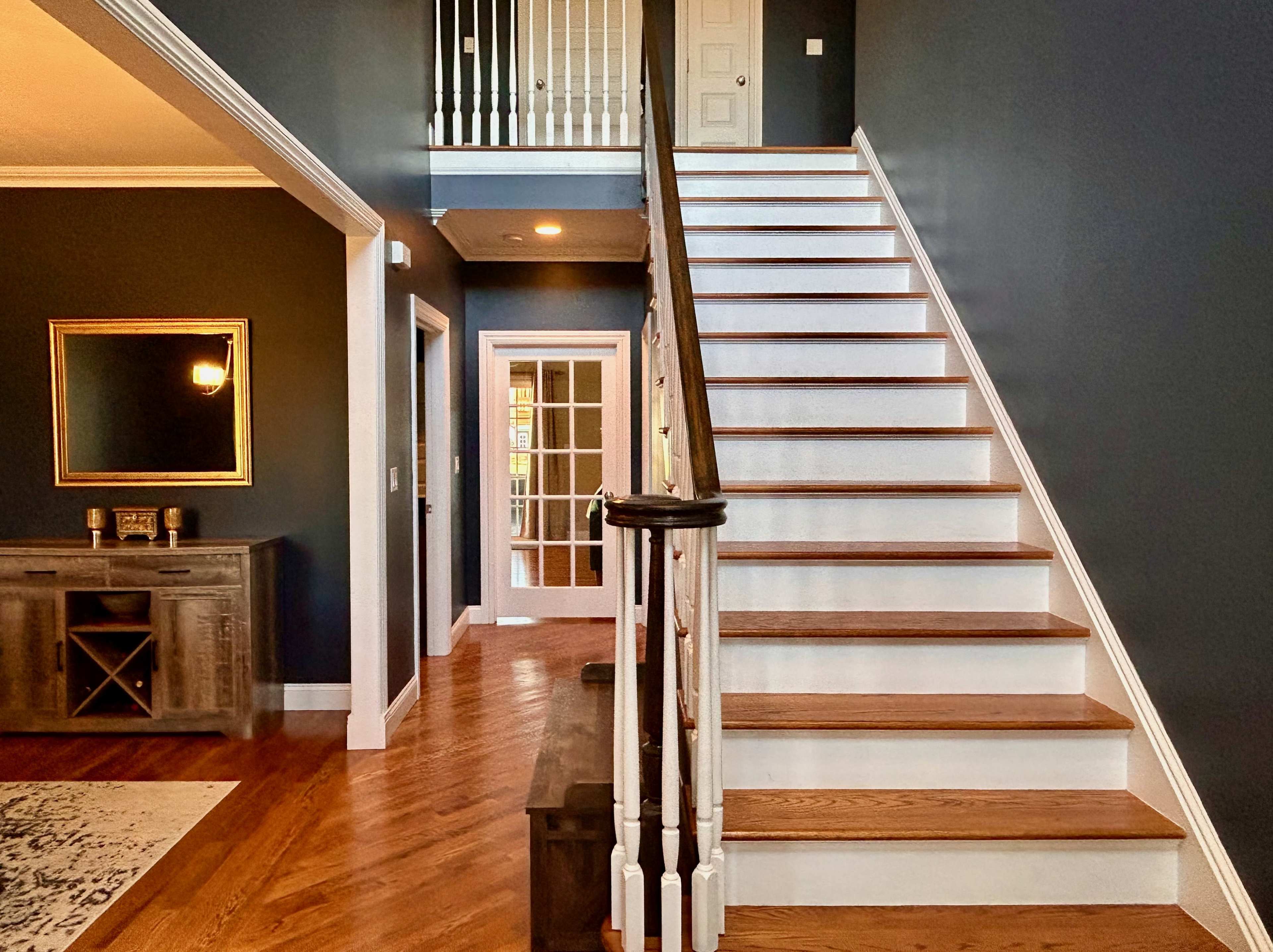A staircase leading to a second floor, with white steps and dark walls, alongside a wooden console table and a doorway in the background.