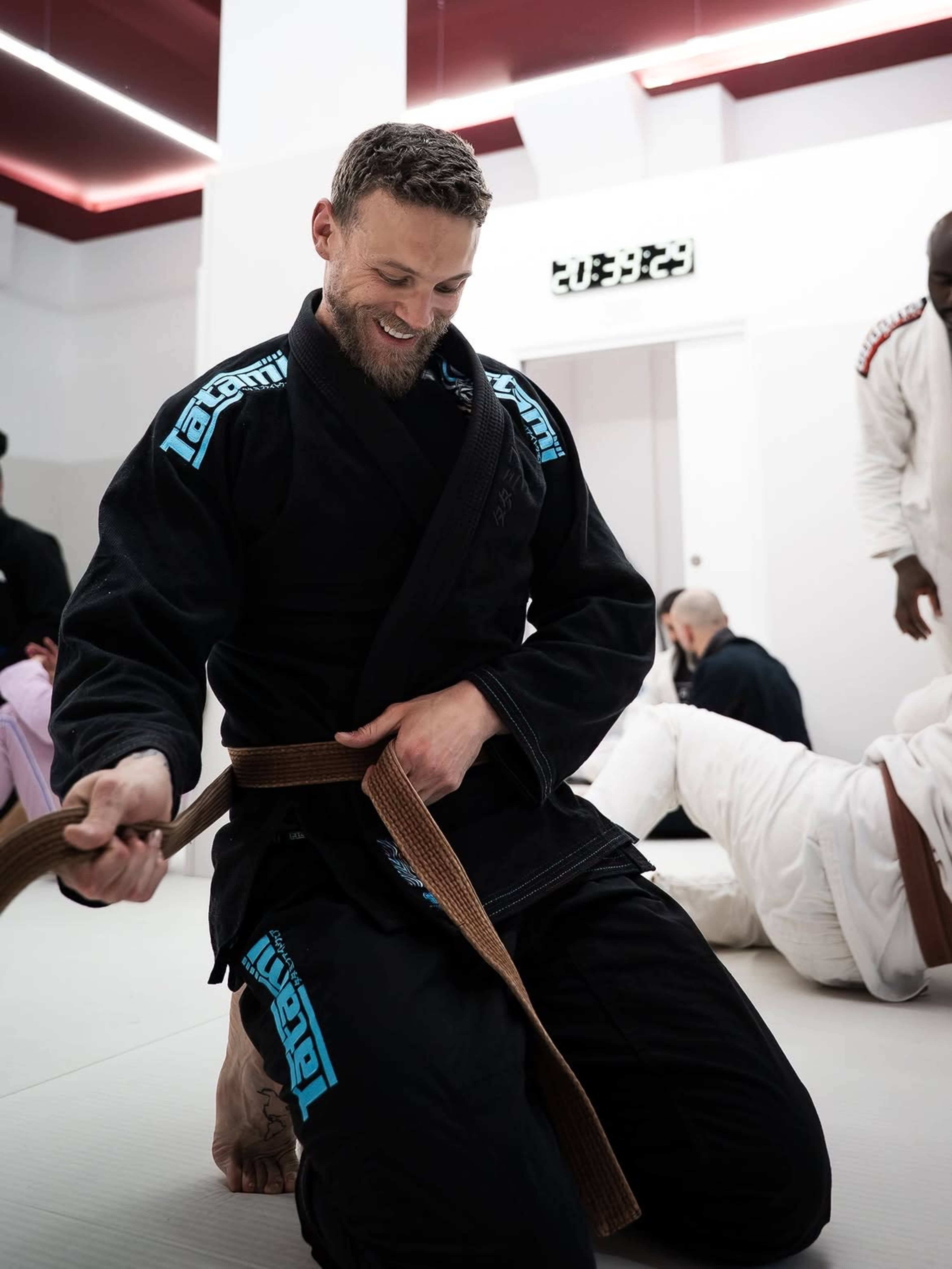 A man in a black jiu-jitsu gi ties his brown belt while kneeling on a mat in a training dojo.