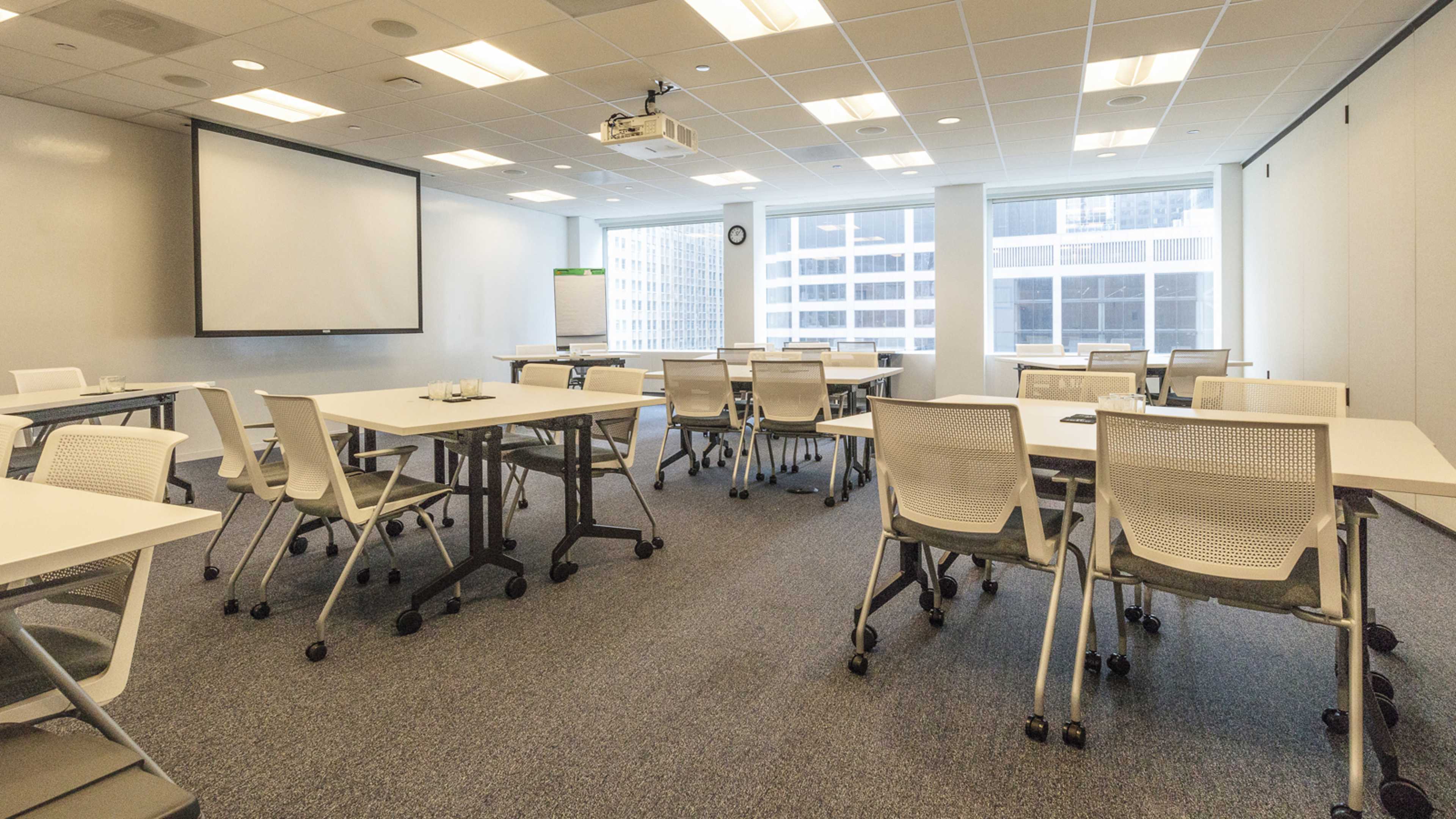 A modern conference room with several movable tables and chairs arranged around a projection screen and large windows.