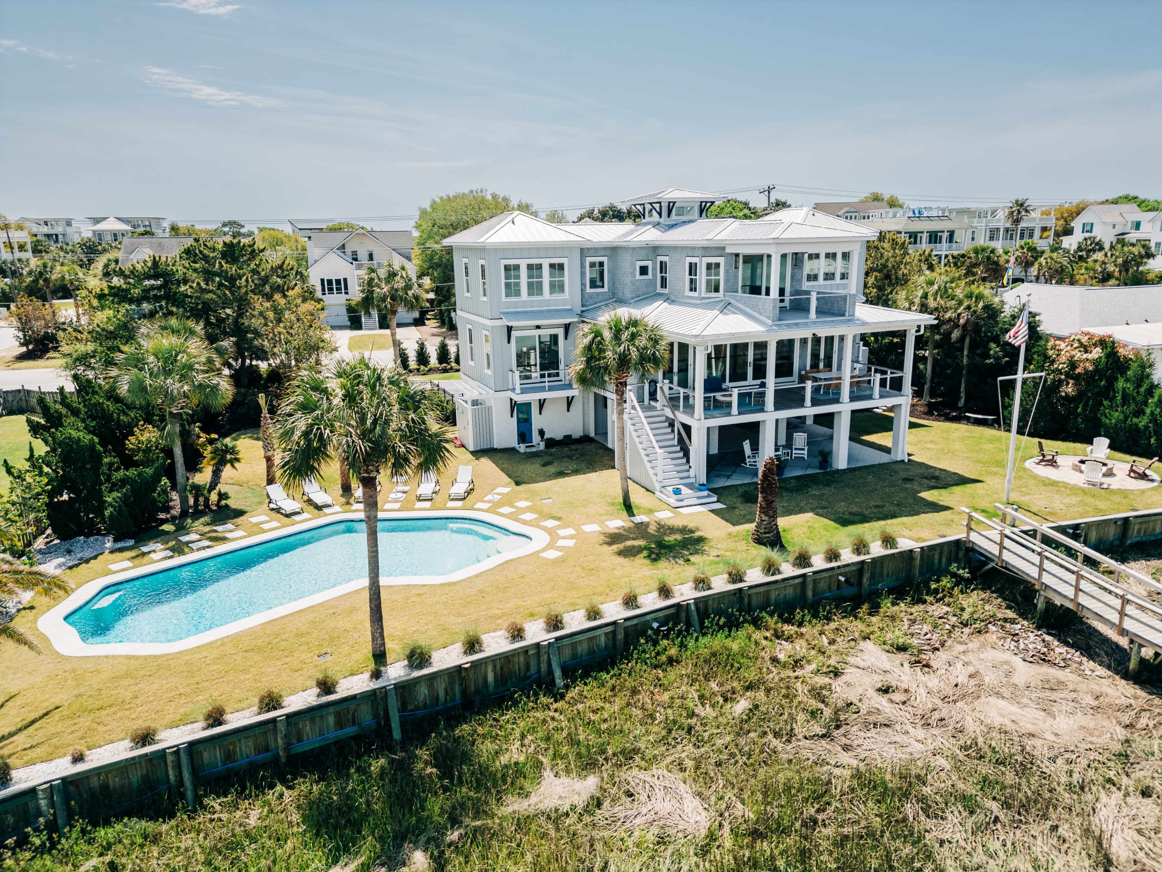 A large coastal house with a swimming pool and palm trees is situated next to a backyard and a wooden walkway leading to the water.
