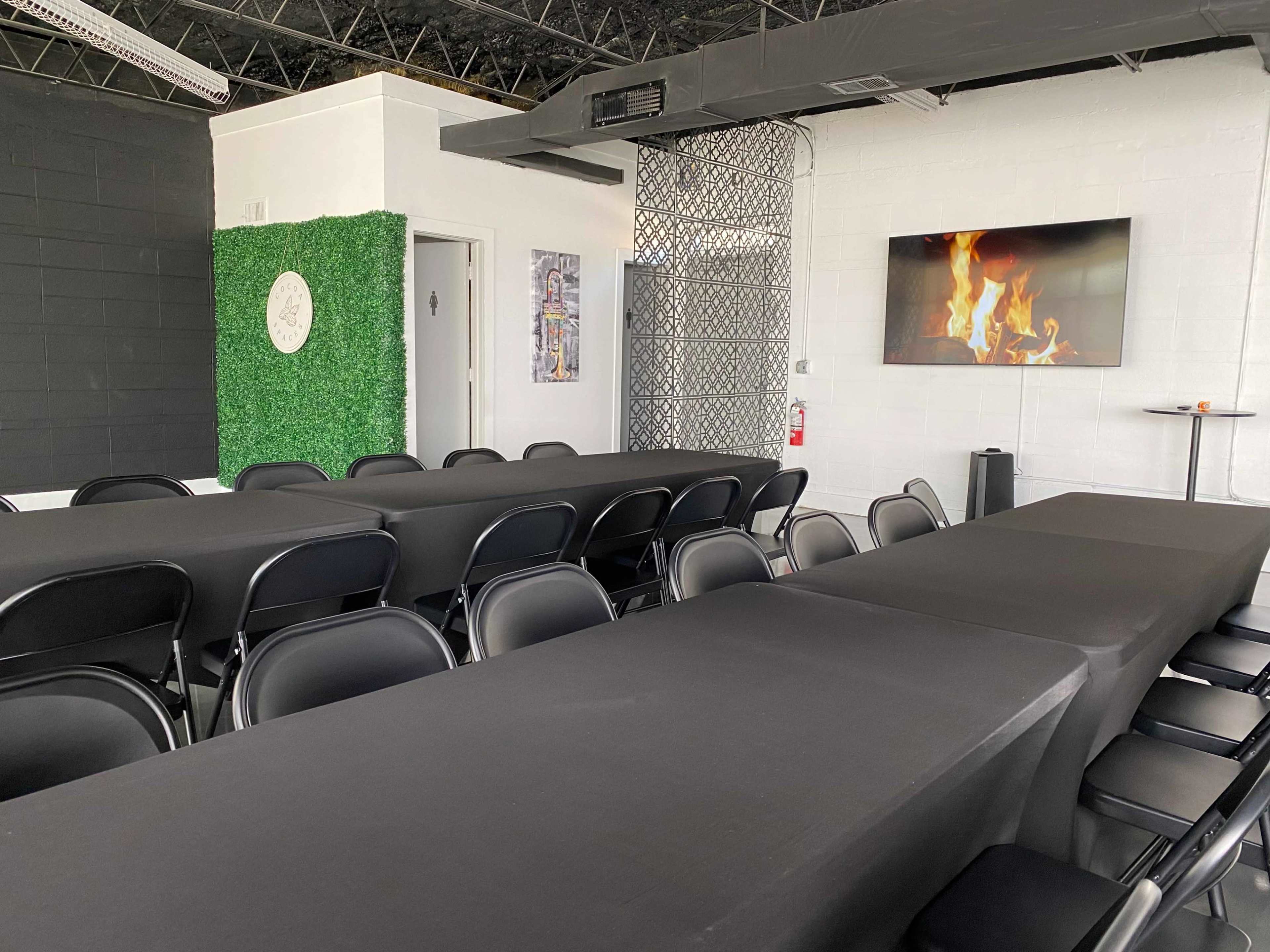 A modern conference room features long black tables with black chairs, a green wall with decorative elements, and a screen displaying a fireplace image.