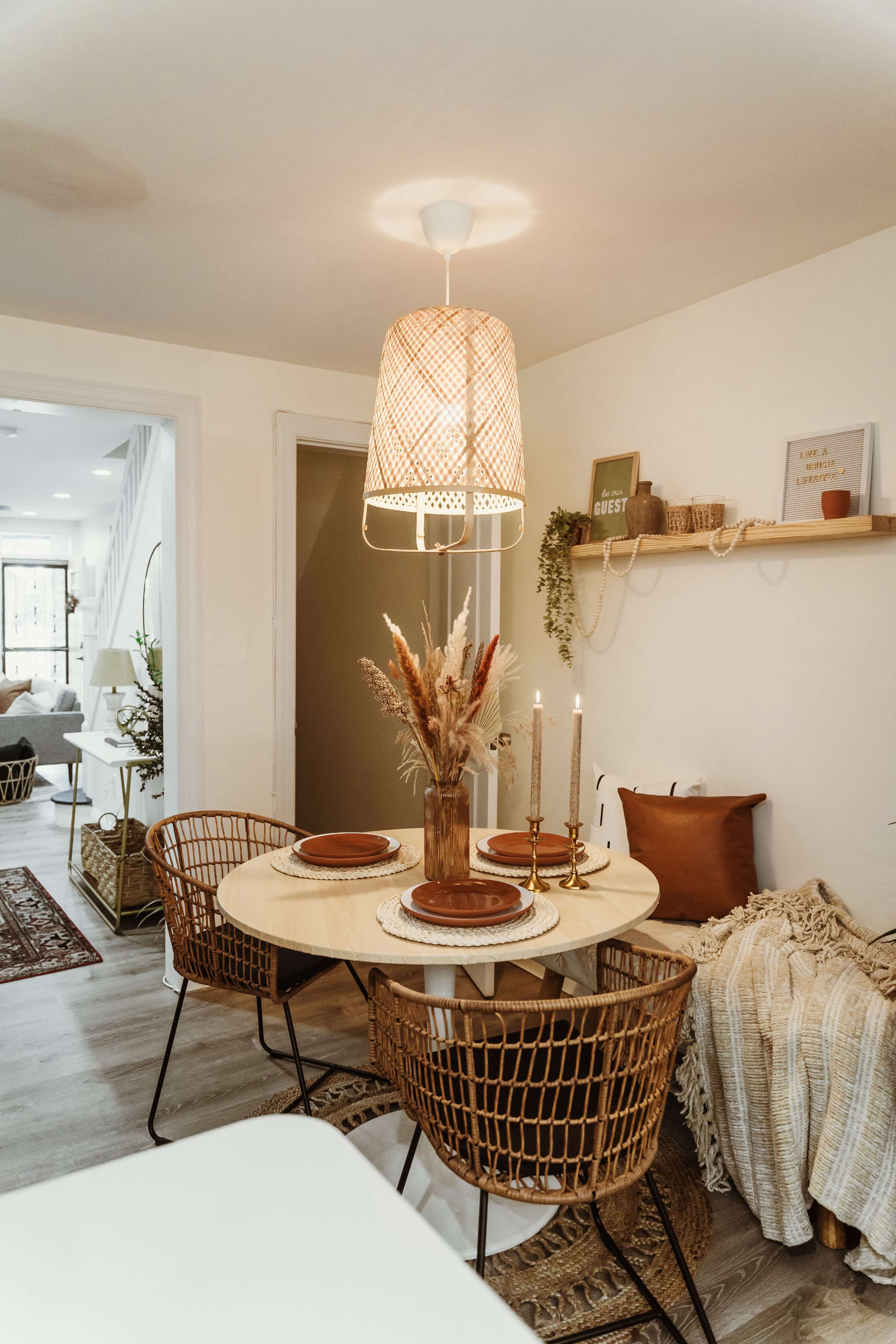 A round dining table is set for two with plates and candles, surrounded by woven chairs in a well-lit, minimally decorated dining area.