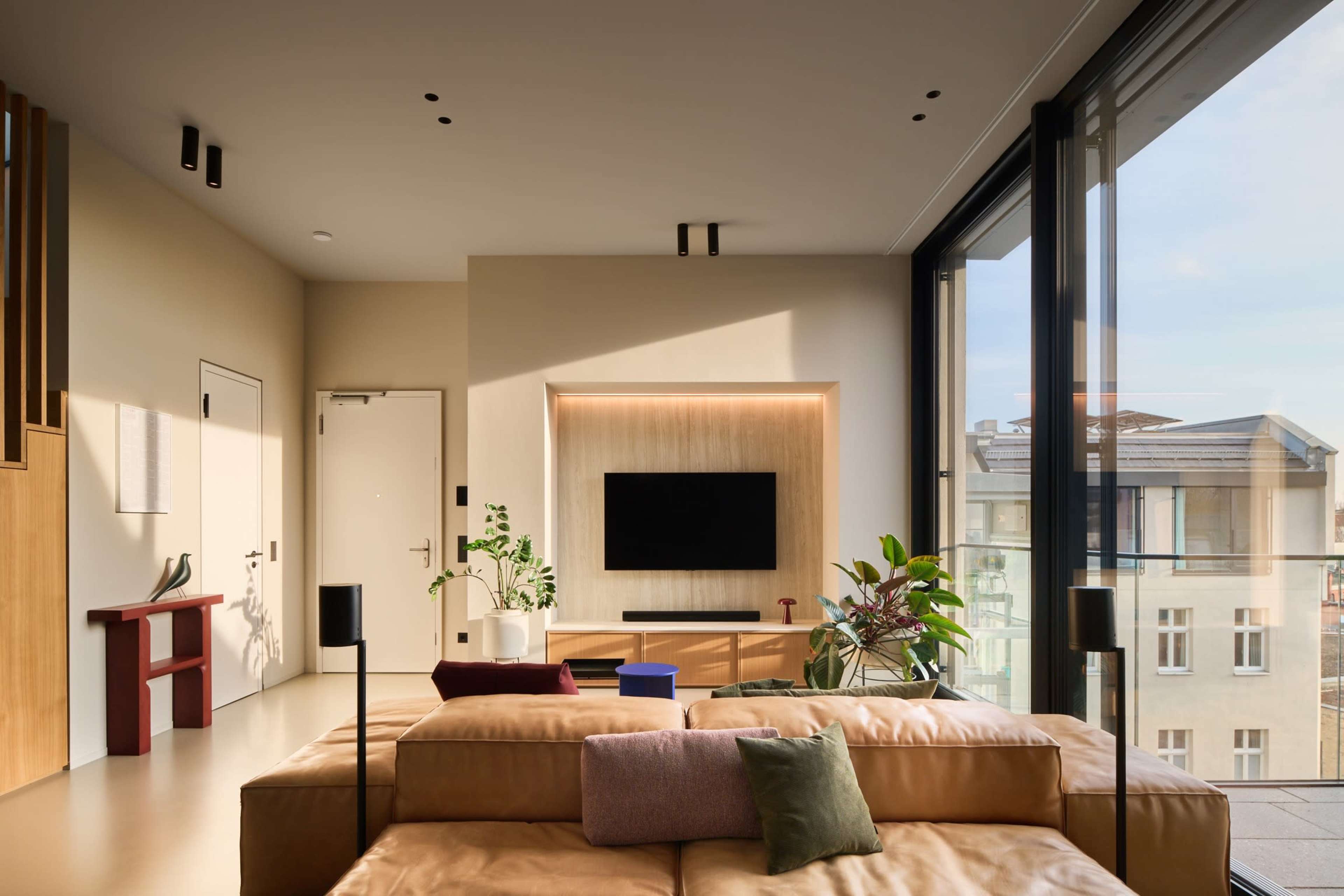 The image shows a modern living room with a large window, featuring a beige sofa, a wall-mounted TV, and various potted plants.