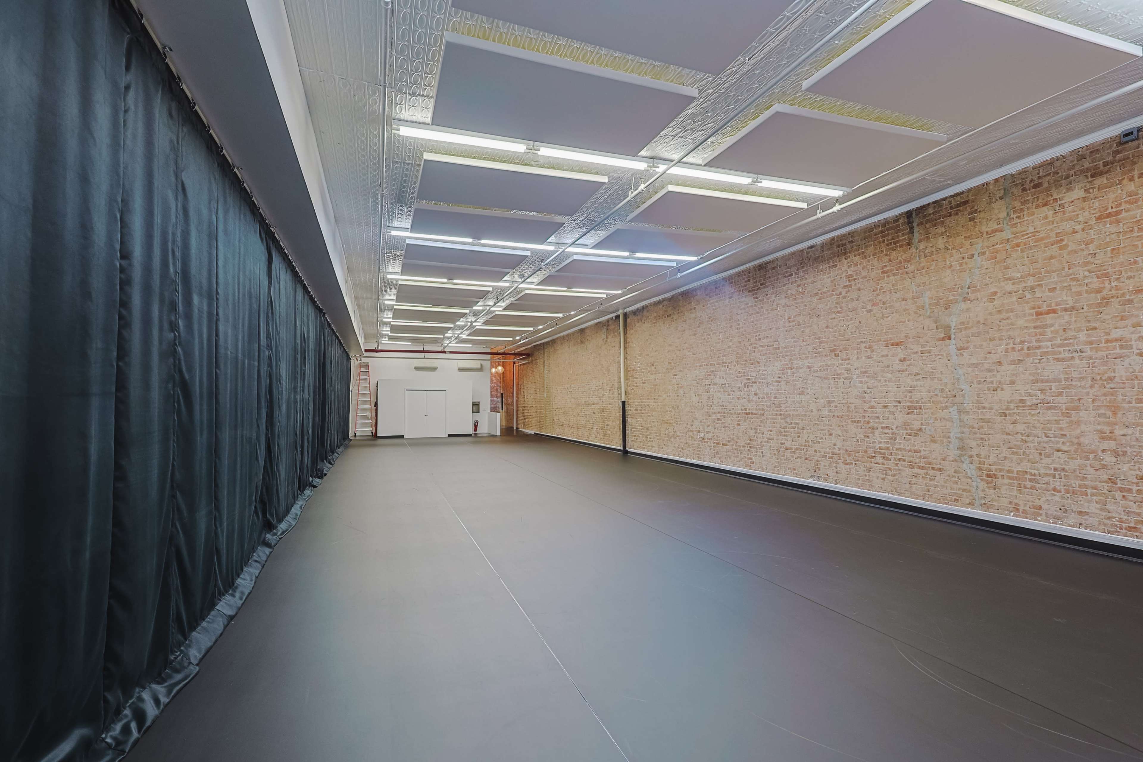 The image shows a spacious interior room with exposed brick walls and a dark floor, featuring overhead lighting and a black curtain along one side.