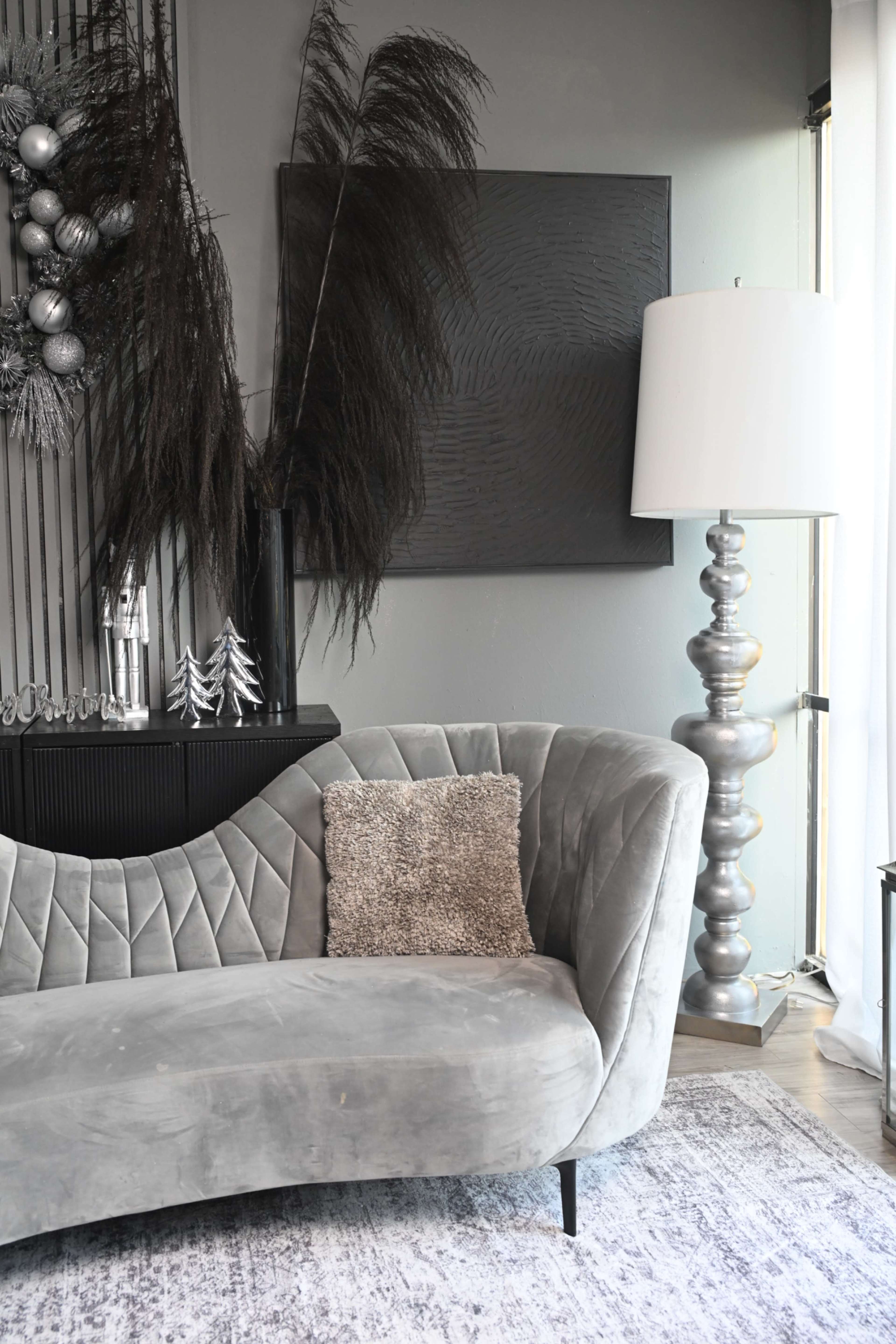 The image features a modern living room setup with a gray velvet curved sofa, decorative wall accents, and a tall lamp beside it.