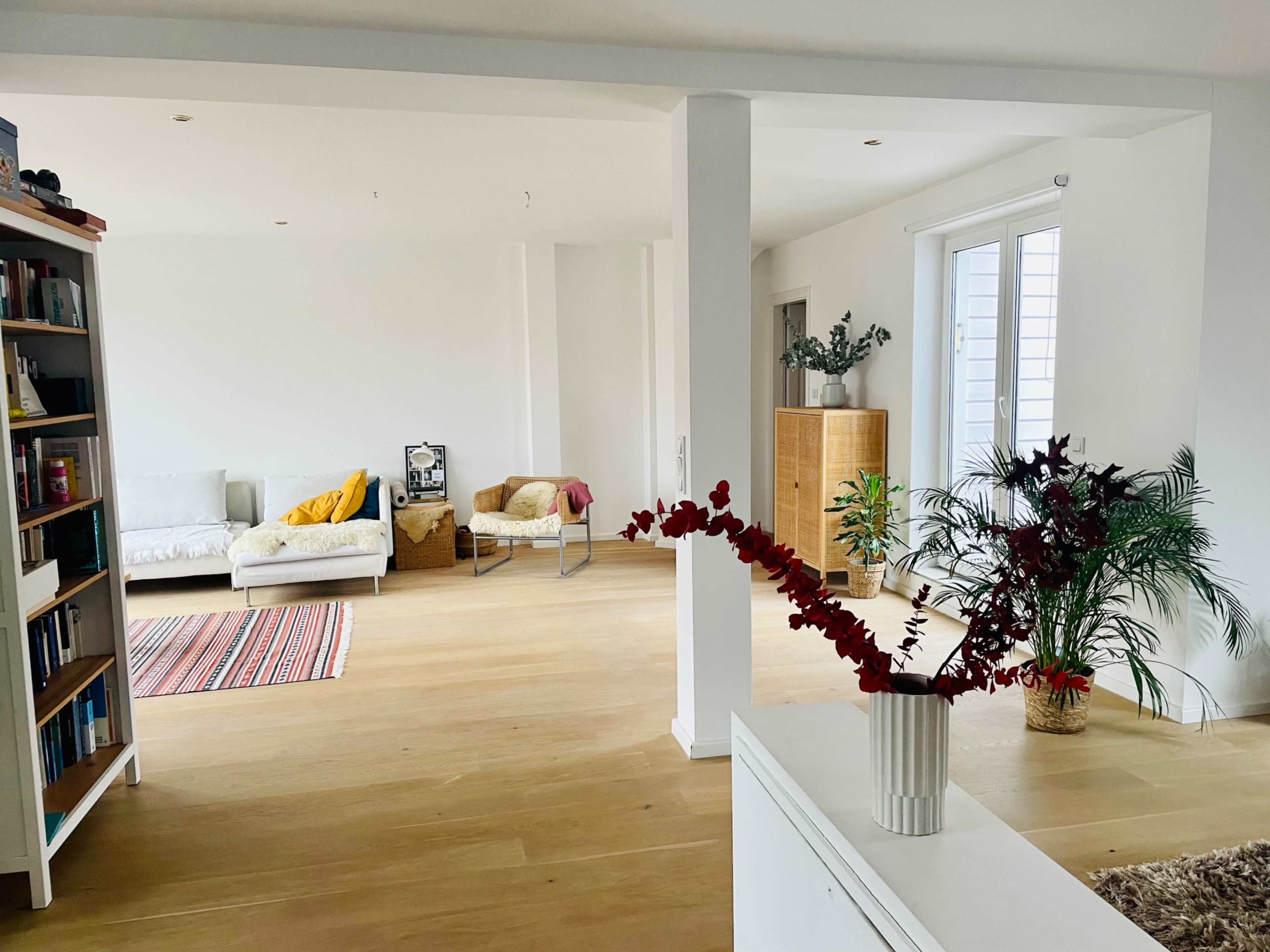 A spacious, minimalist living area with a wooden floor, a bookshelf, and various plants.