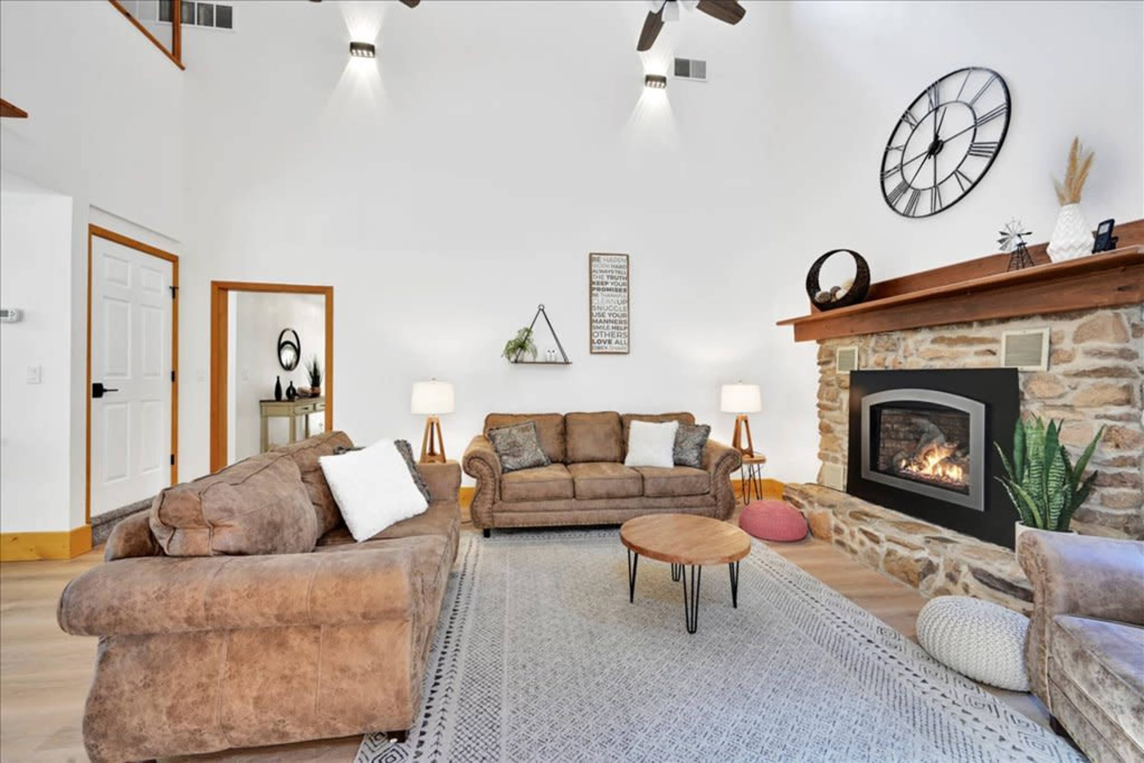A living room with two brown sofas, a round coffee table, and a stone fireplace beneath a large clock on the wall.