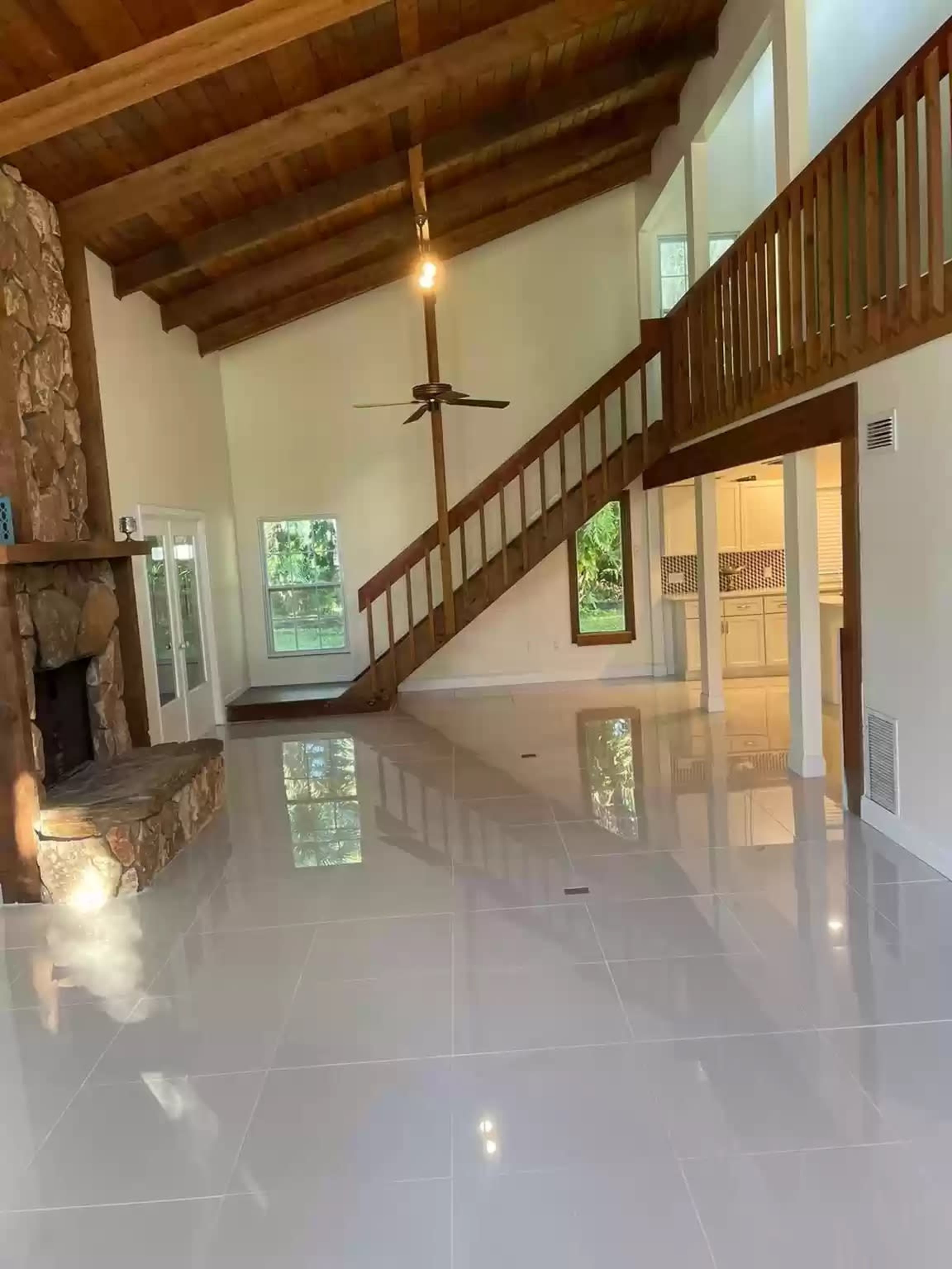 A spacious living area with a stone fireplace, high wooden beam ceiling, and a staircase leading to an upper level, all featuring polished white tile flooring.