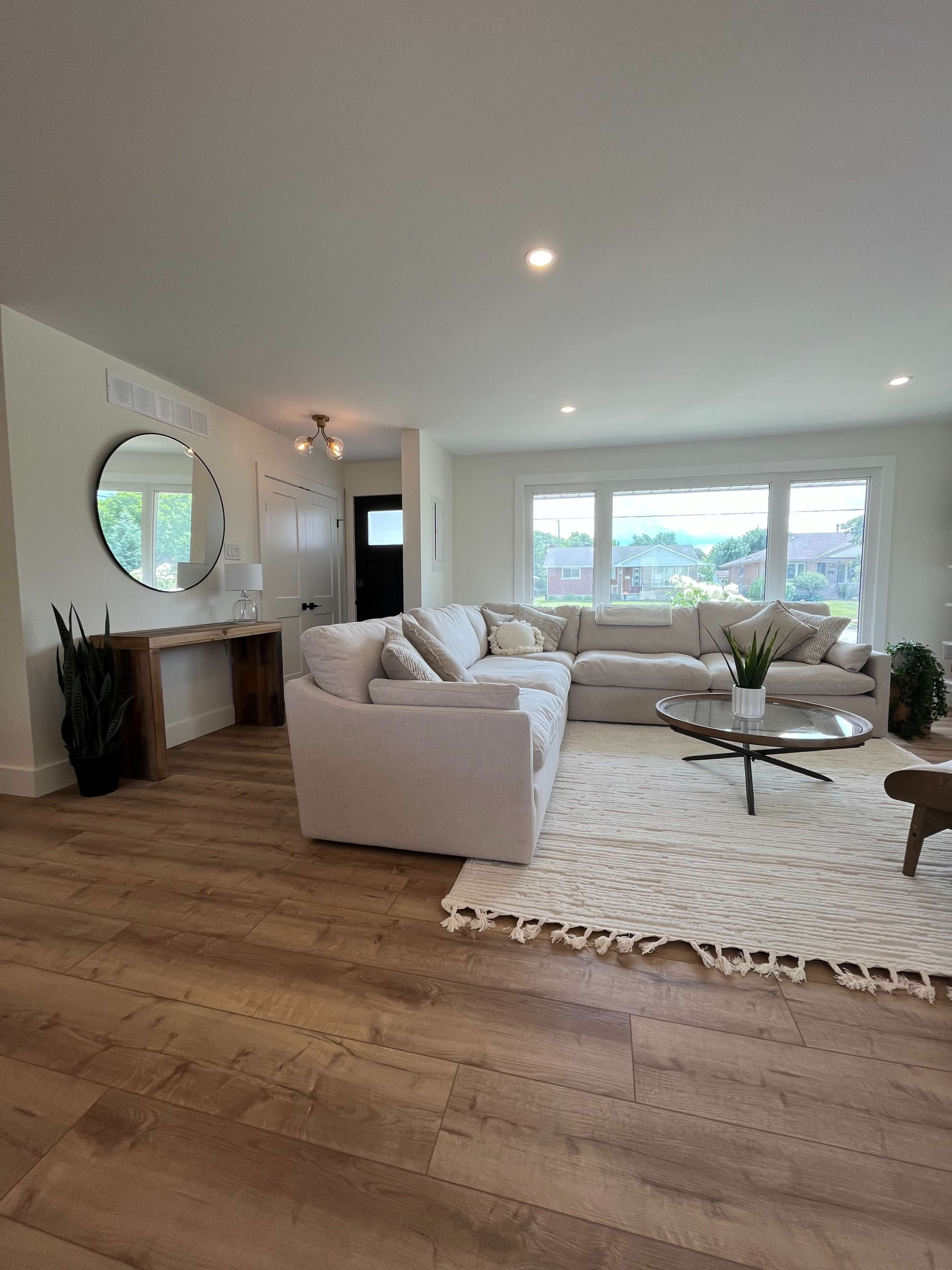 A modern living room featuring a large light-colored sectional couch, a round coffee table, and a rug on a wooden floor, with large windows allowing natural light to enter.