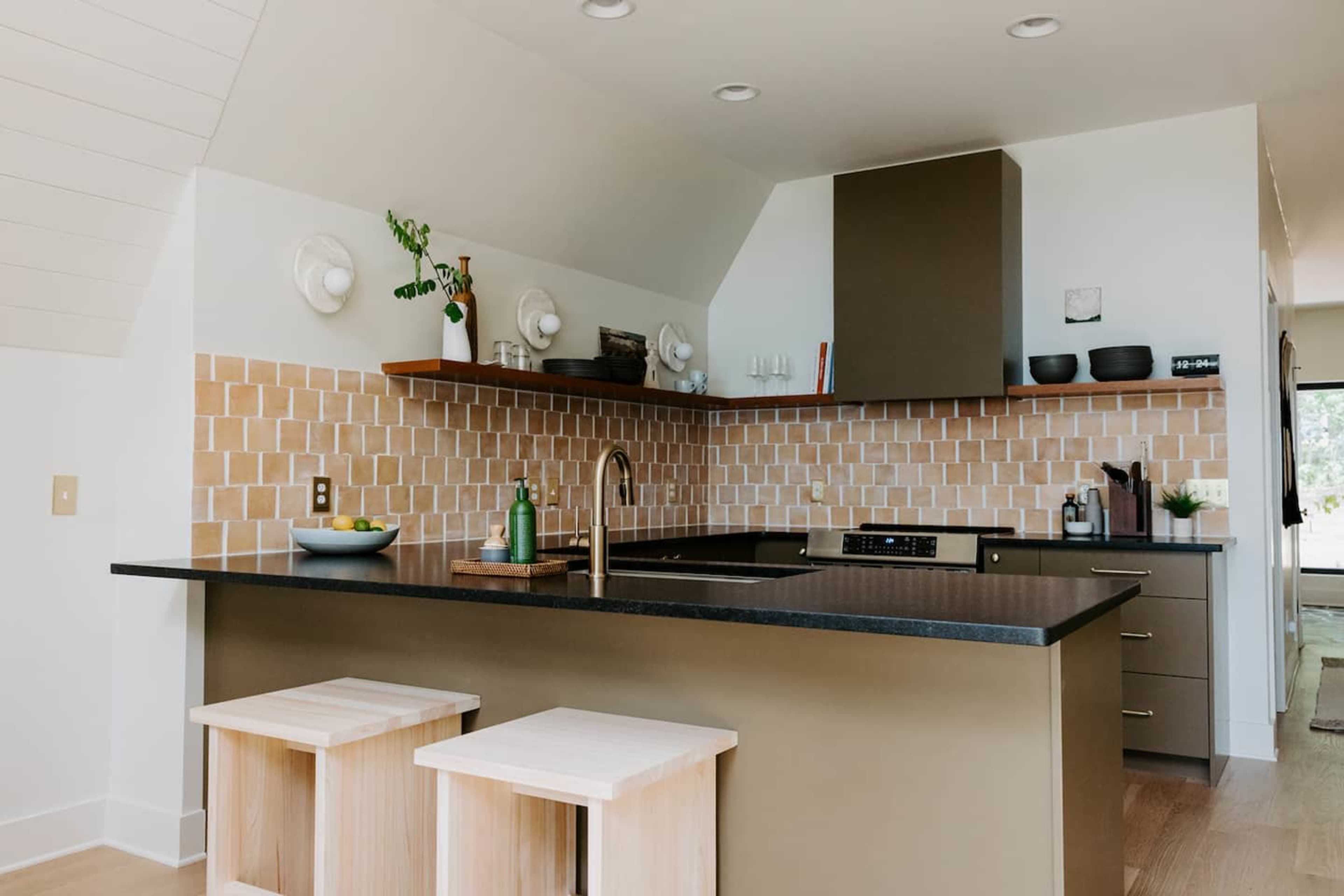 The image shows a modern kitchen with a dark countertop, light-colored tiled backsplash, and minimalist wooden stools.