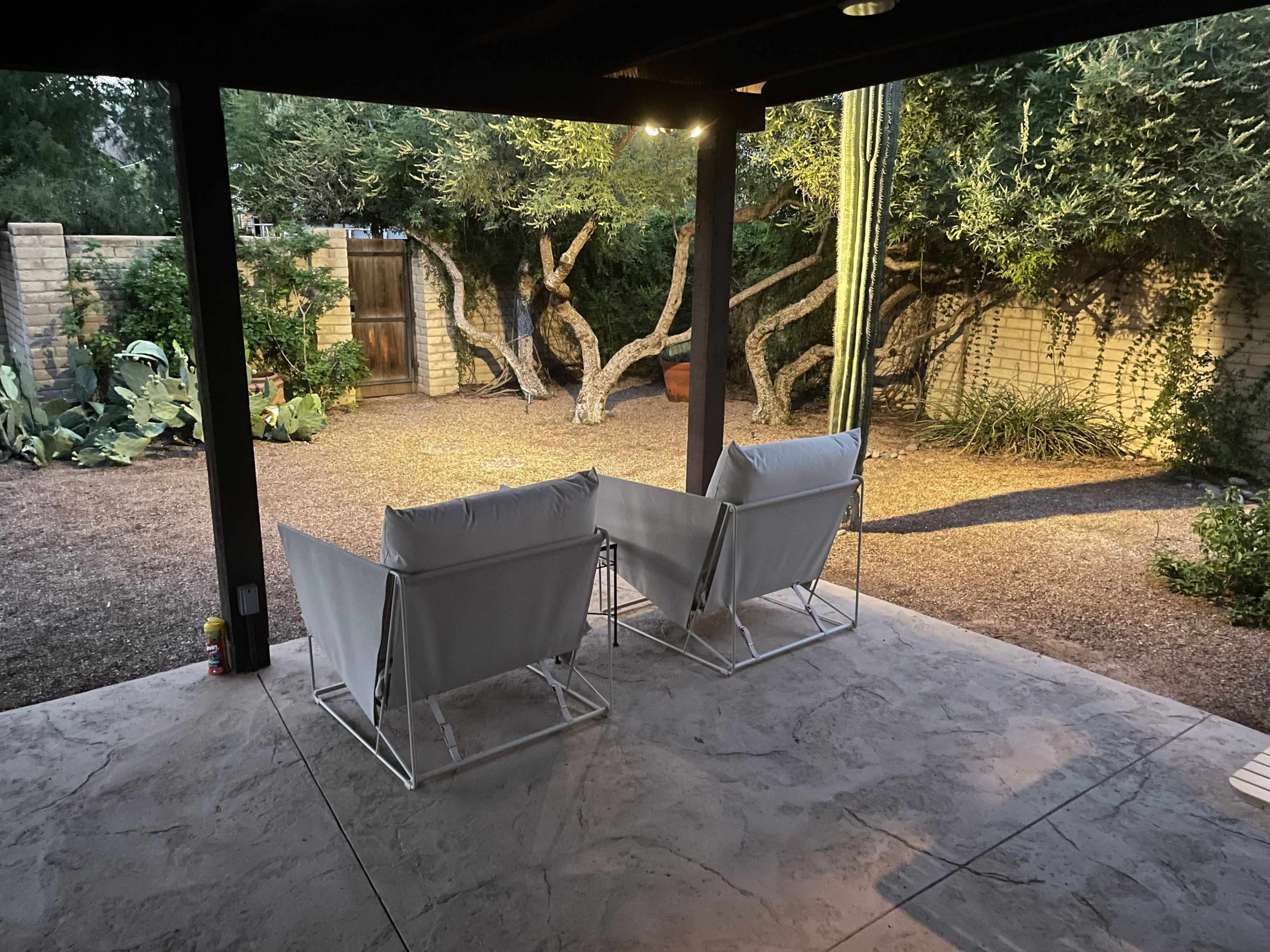 Two white folding chairs are positioned in a shaded patio area, overlooking a gravel garden with various plants and a tall cactus.