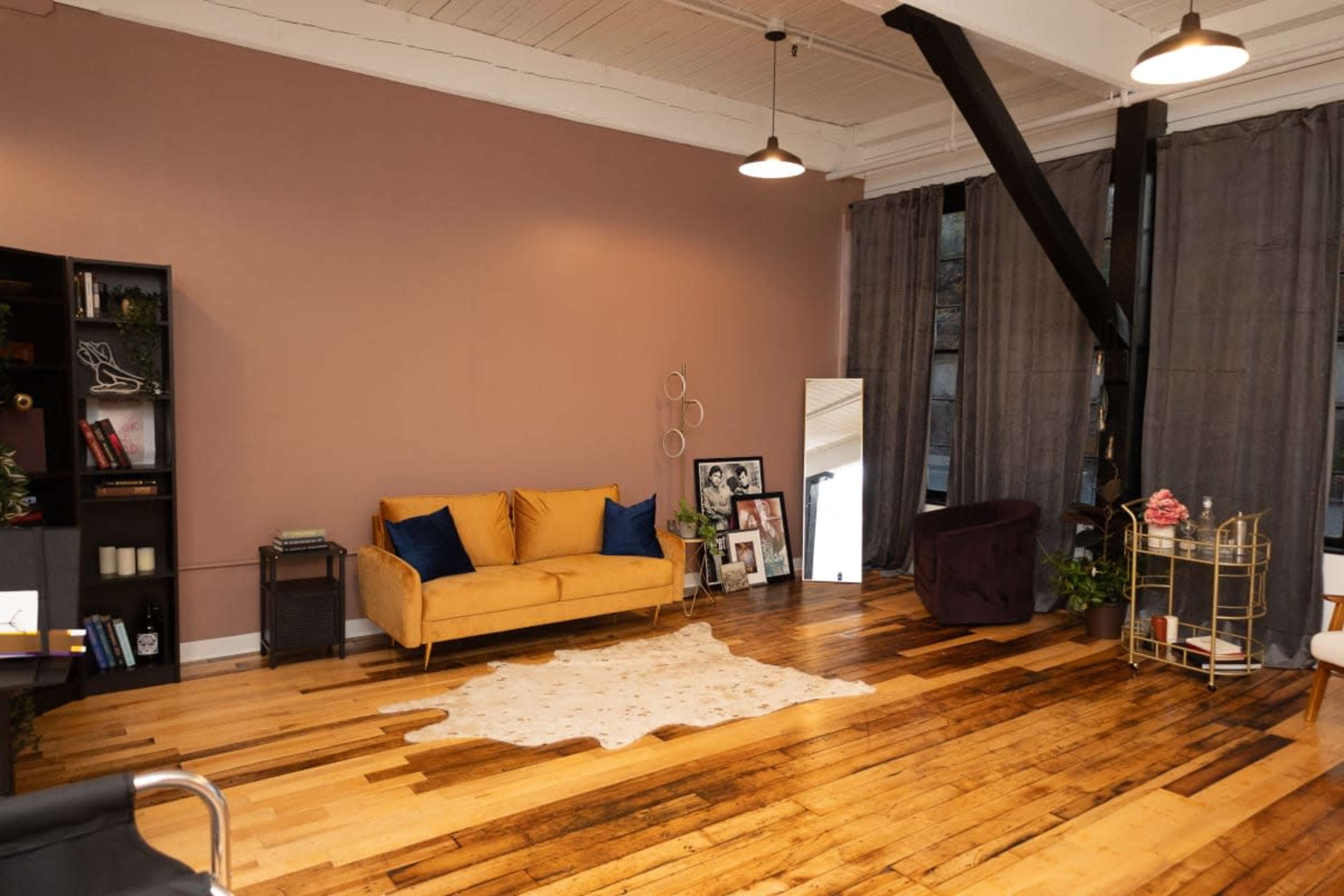 A spacious interior with hardwood floors, featuring a yellow sofa, a dark armchair, a decorative mirror, and various plants.