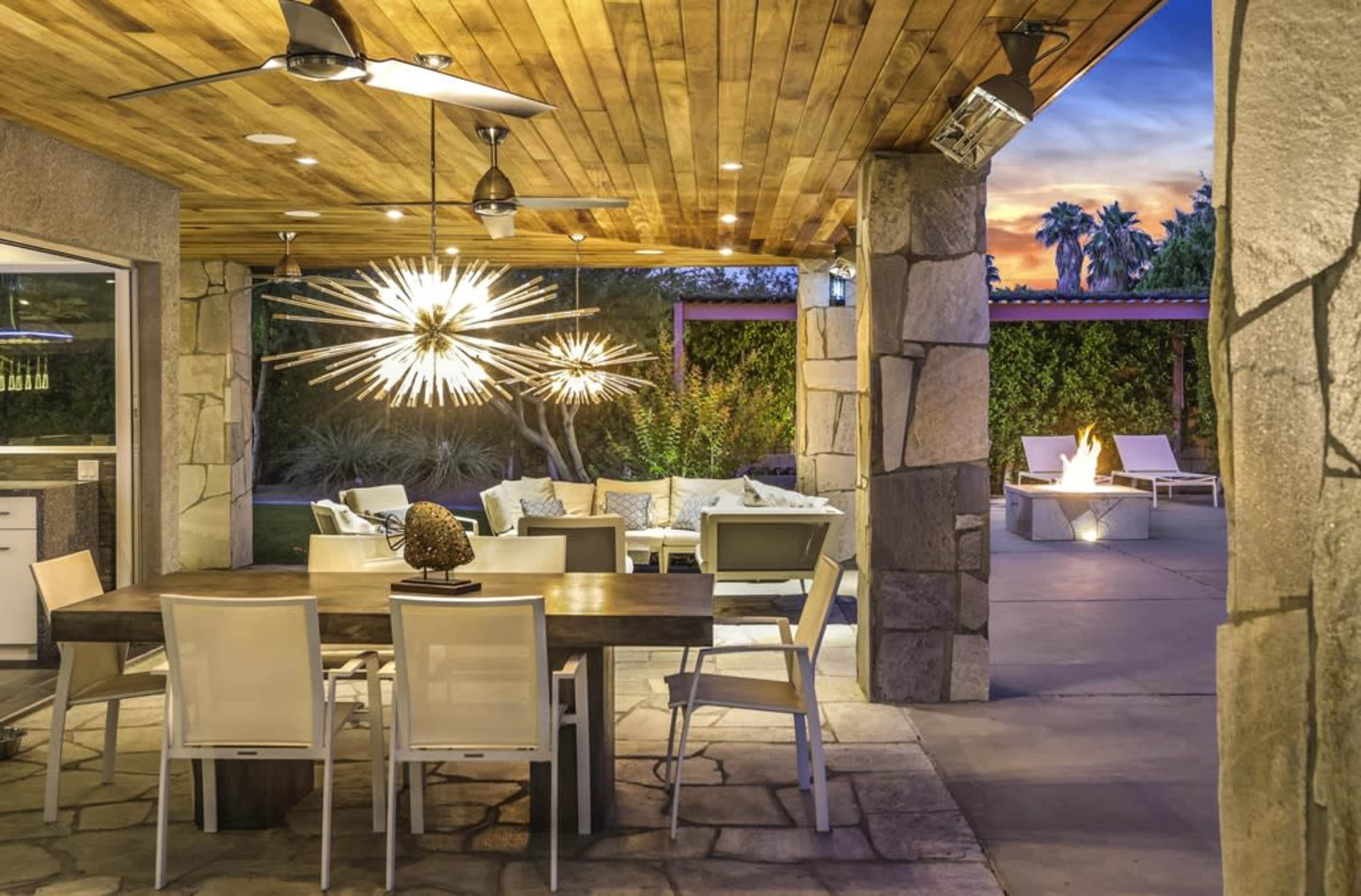 The image shows a stylish outdoor patio area with a dining table, seating arrangement, and a fire pit, surrounded by greenery and illuminated by pendant lights.