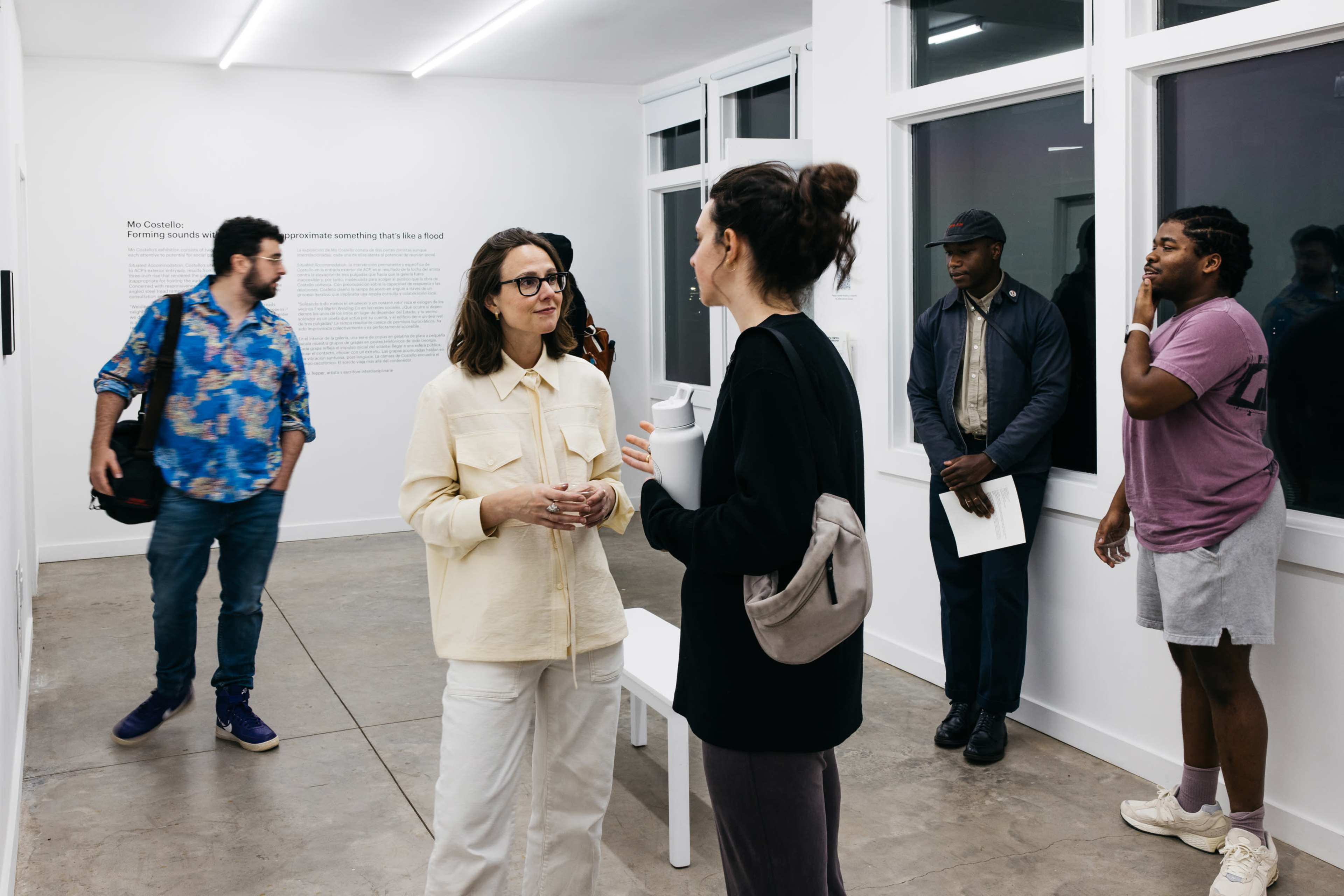 A group of people is gathered in a modern art gallery, with two individuals engaged in conversation at the forefront.