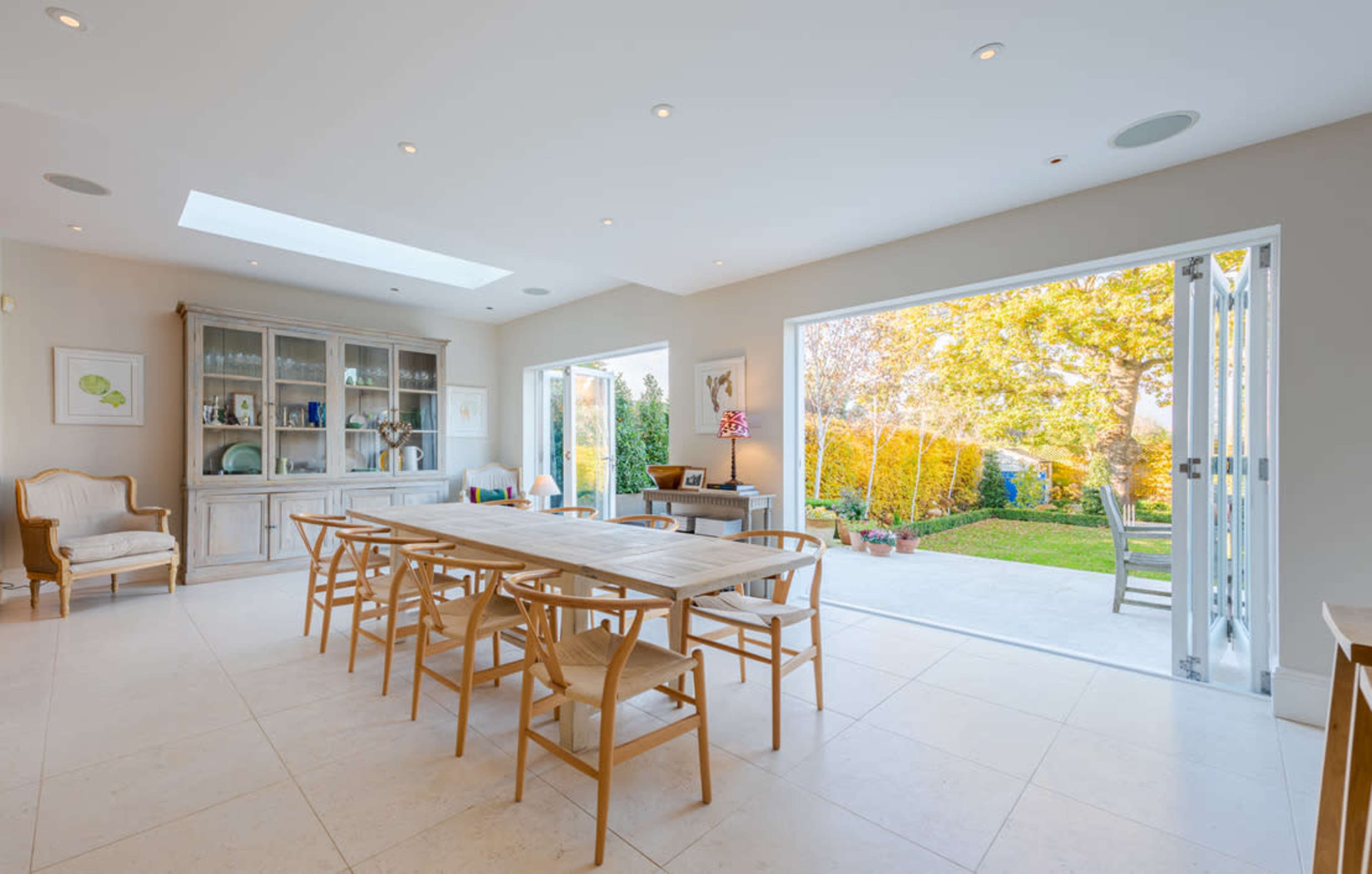A spacious dining room features a long wooden table surrounded by chairs, with large windows opening to a garden outside.