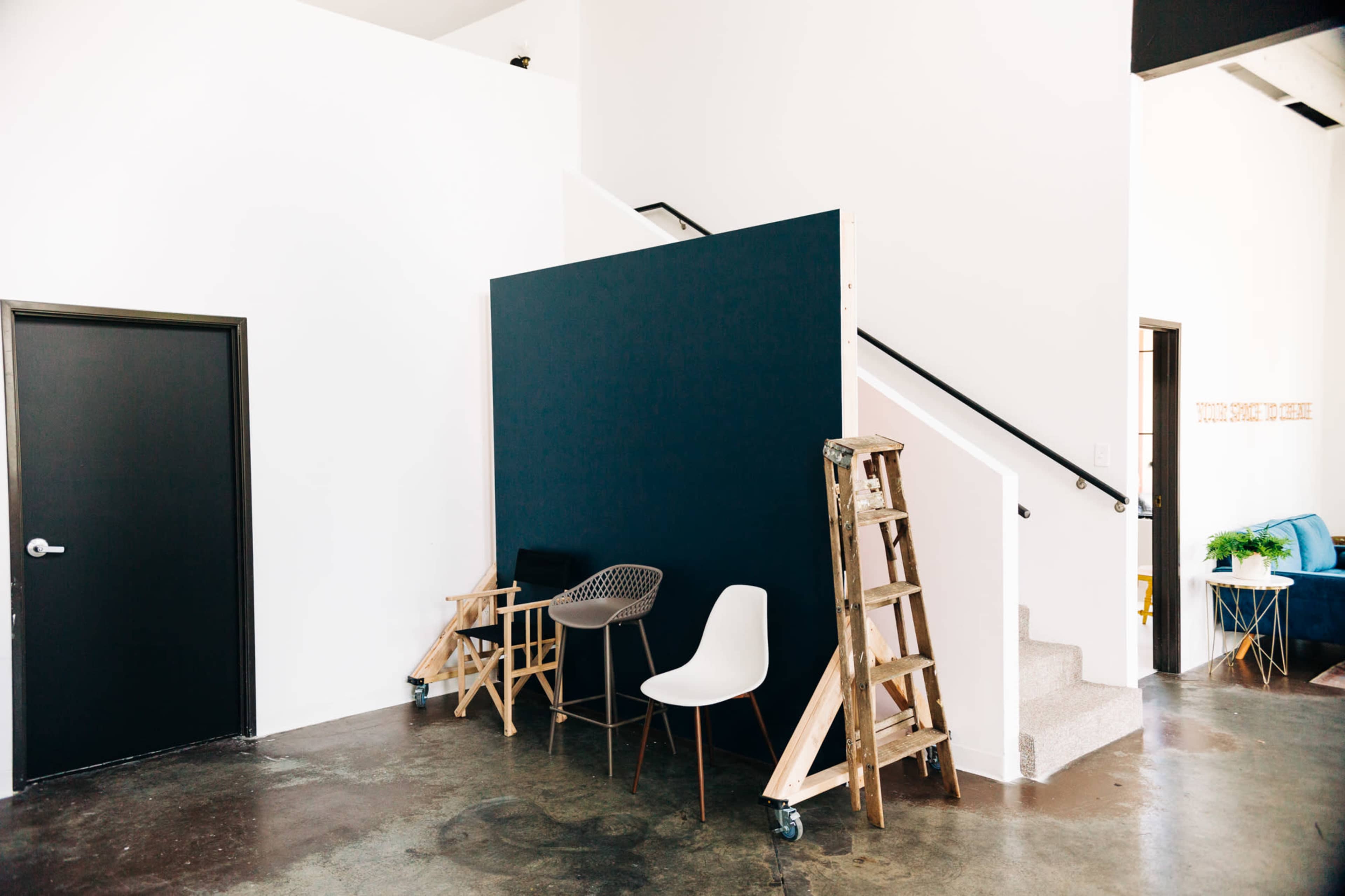 A minimalist indoor space featuring a black wall section, two chairs, a wooden ladder, and a staircase in the background.
