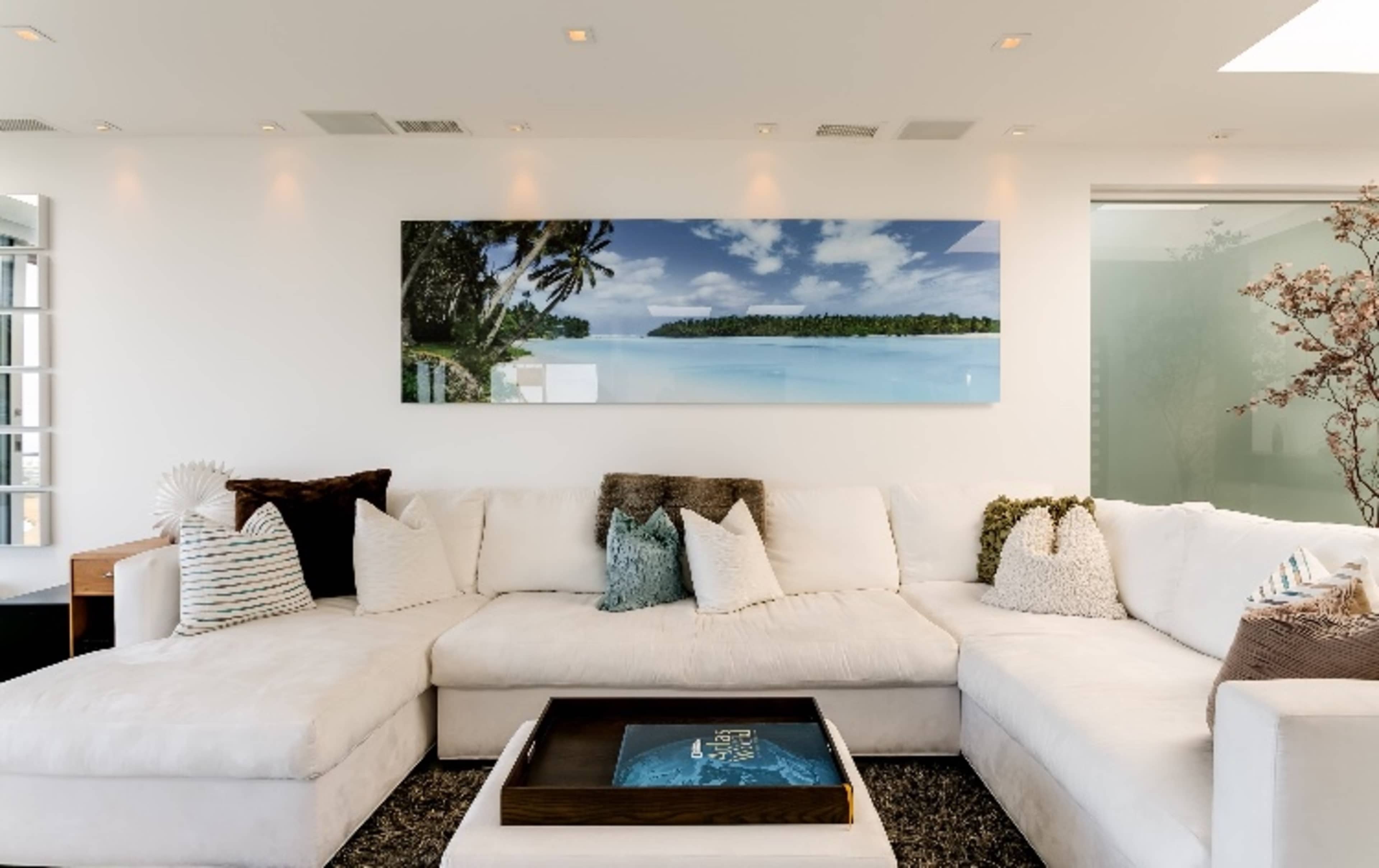 A spacious living room features a large sectional sofa adorned with various throw pillows and a coffee table, with a panoramic artwork of a tropical beach on the wall.