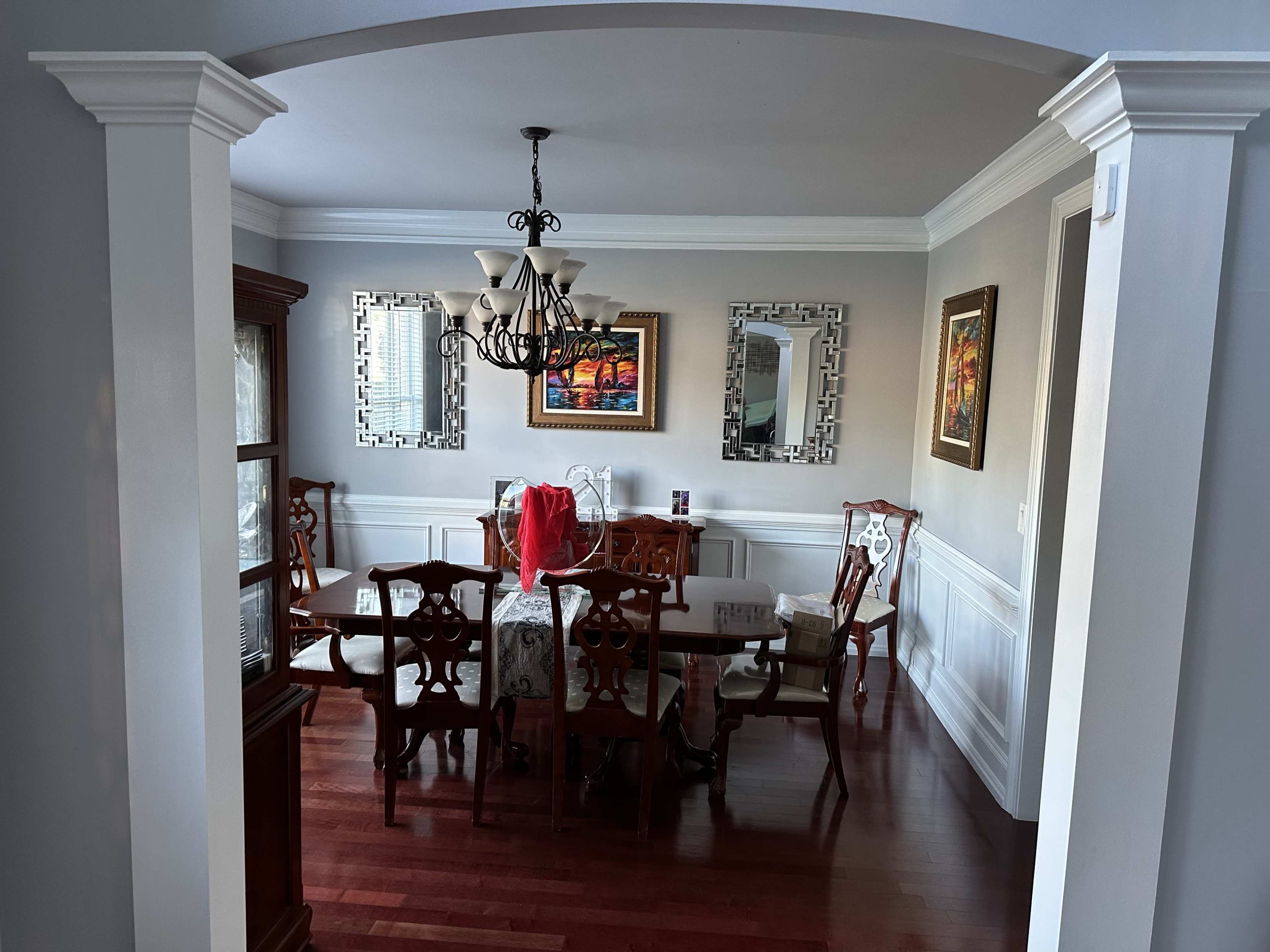The image shows a dining room with a large wooden table surrounded by chairs, framed mirrors on the walls, and a chandelier overhead.