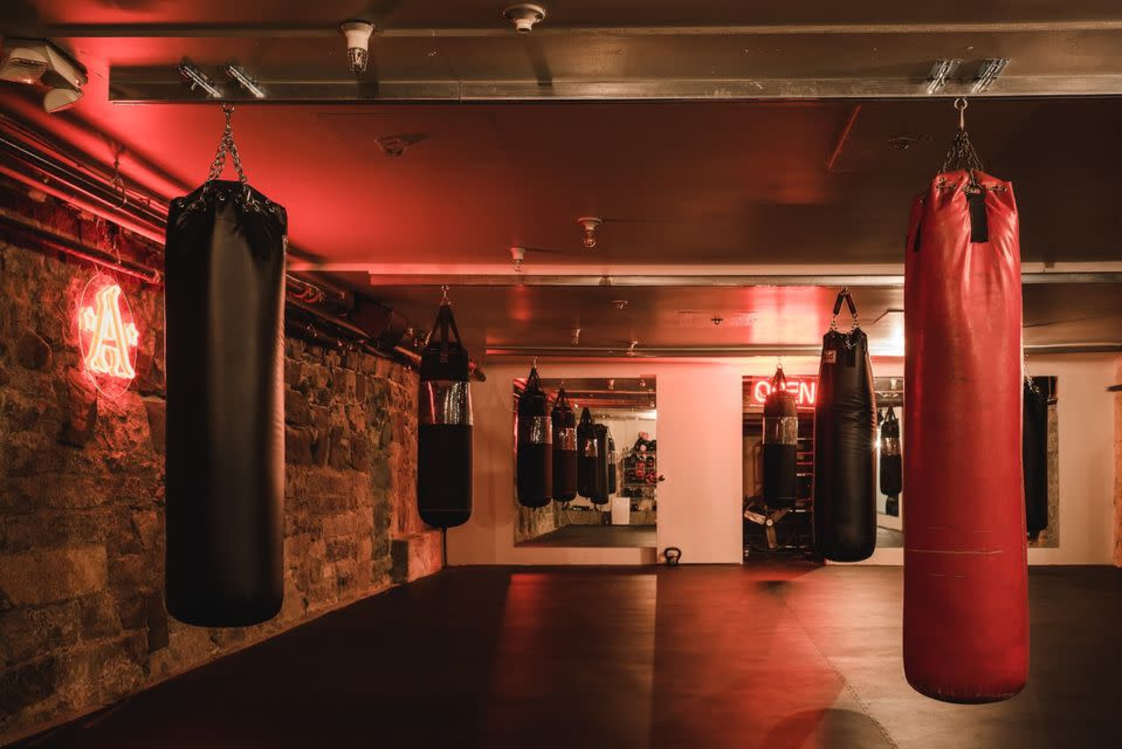 A boxing gym features multiple punching bags hanging from the ceiling, with a neon "OPEN" sign and illuminated walls.