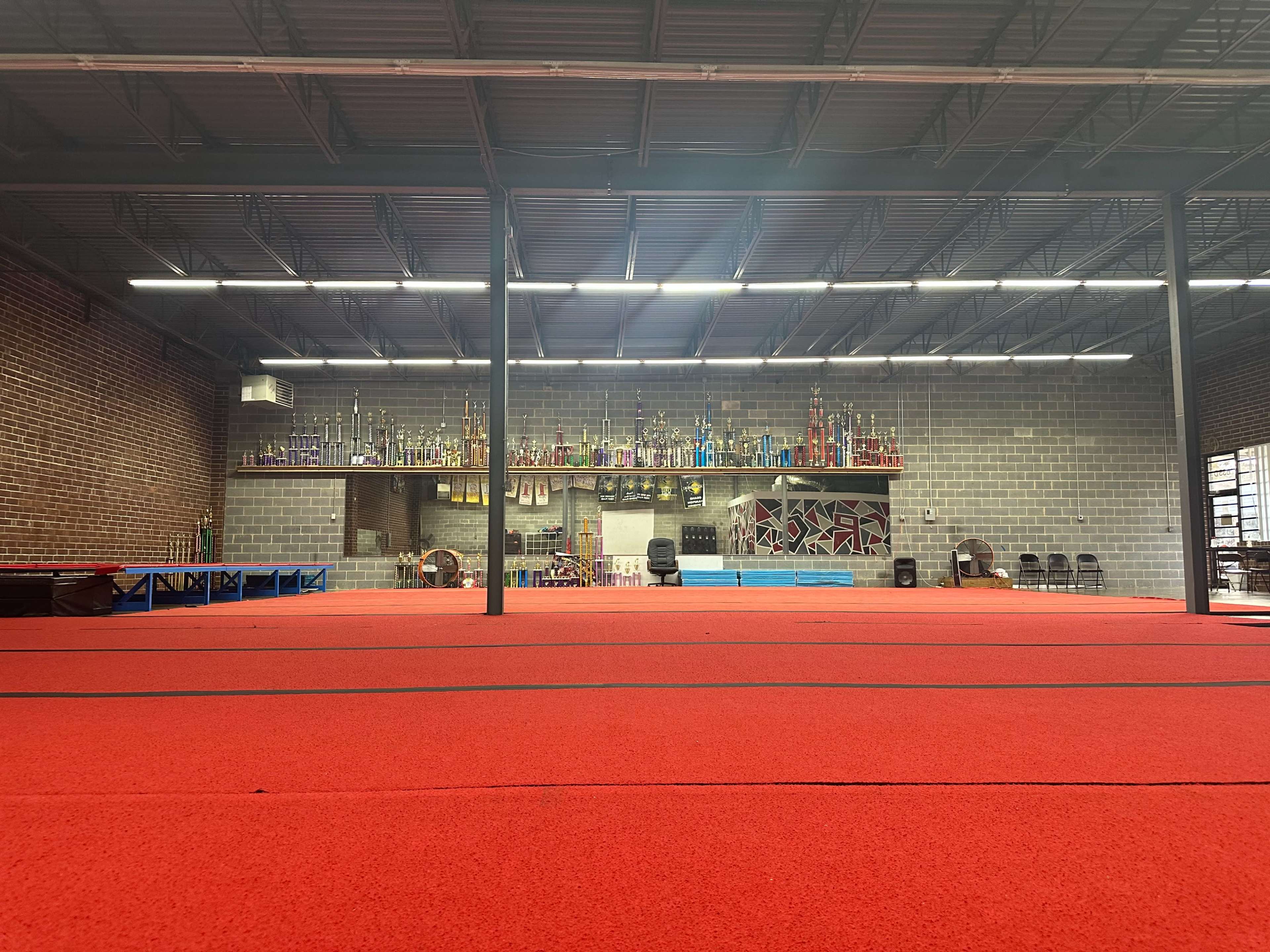 The image shows a gymnasium with a red floor, lined with trophies on shelves against a brick wall, and blue mats in the center.