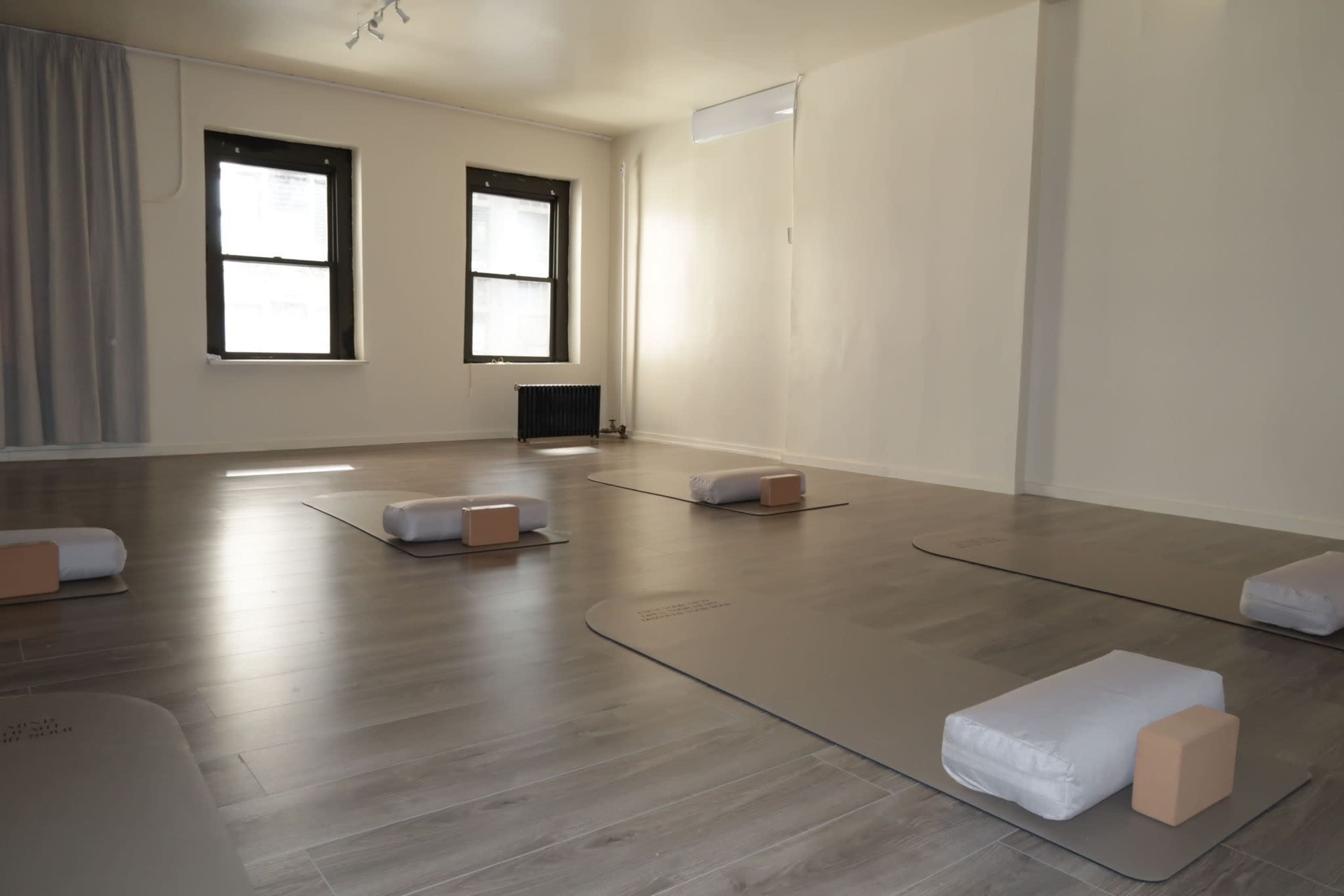 The image shows a spacious yoga studio with four exercise mats and props arranged neatly on a light wooden floor, surrounded by white walls and large windows.