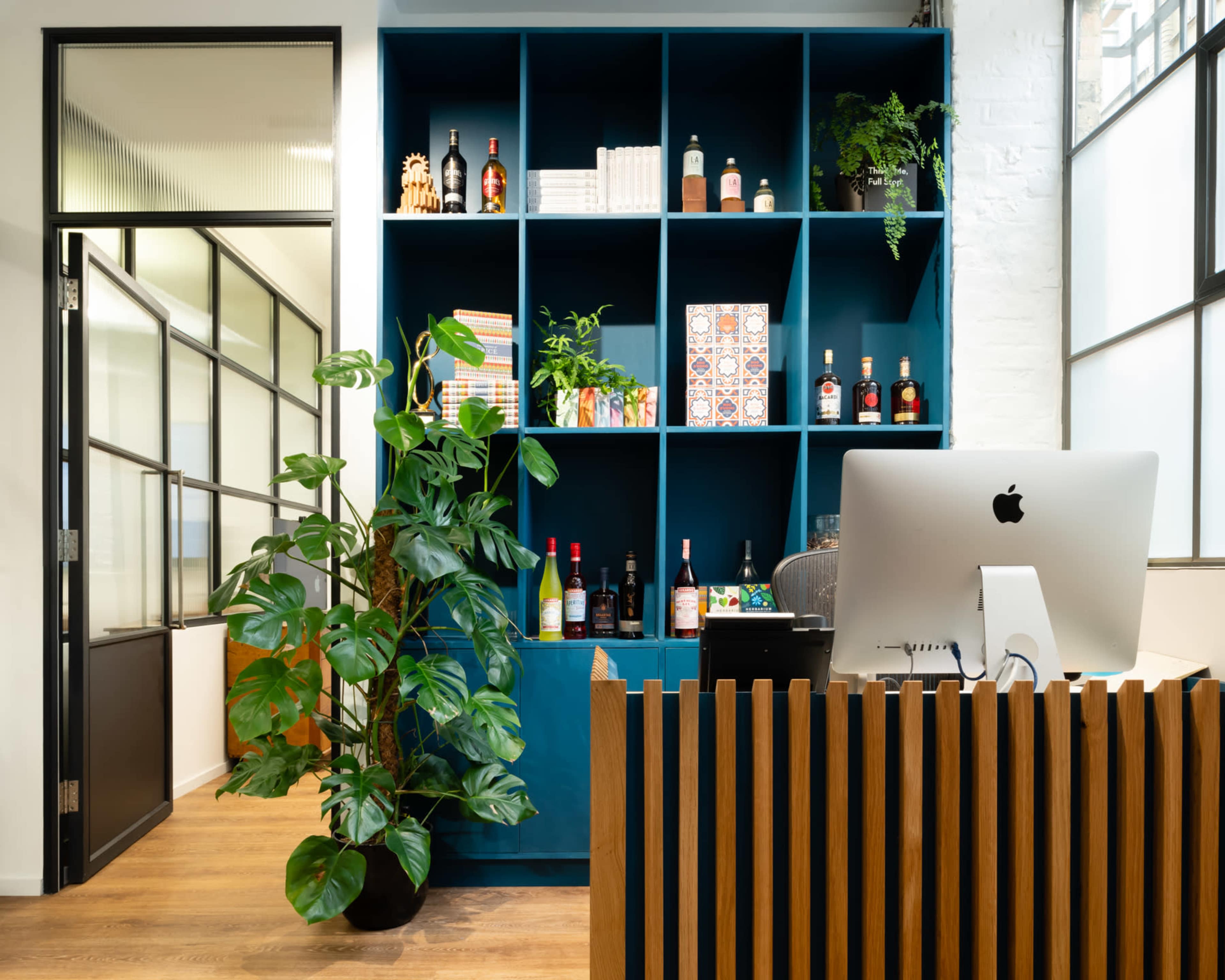 A modern office space with a blue shelving unit displaying bottles and decorative items, a potted plant, and a reception desk featuring an iMac.