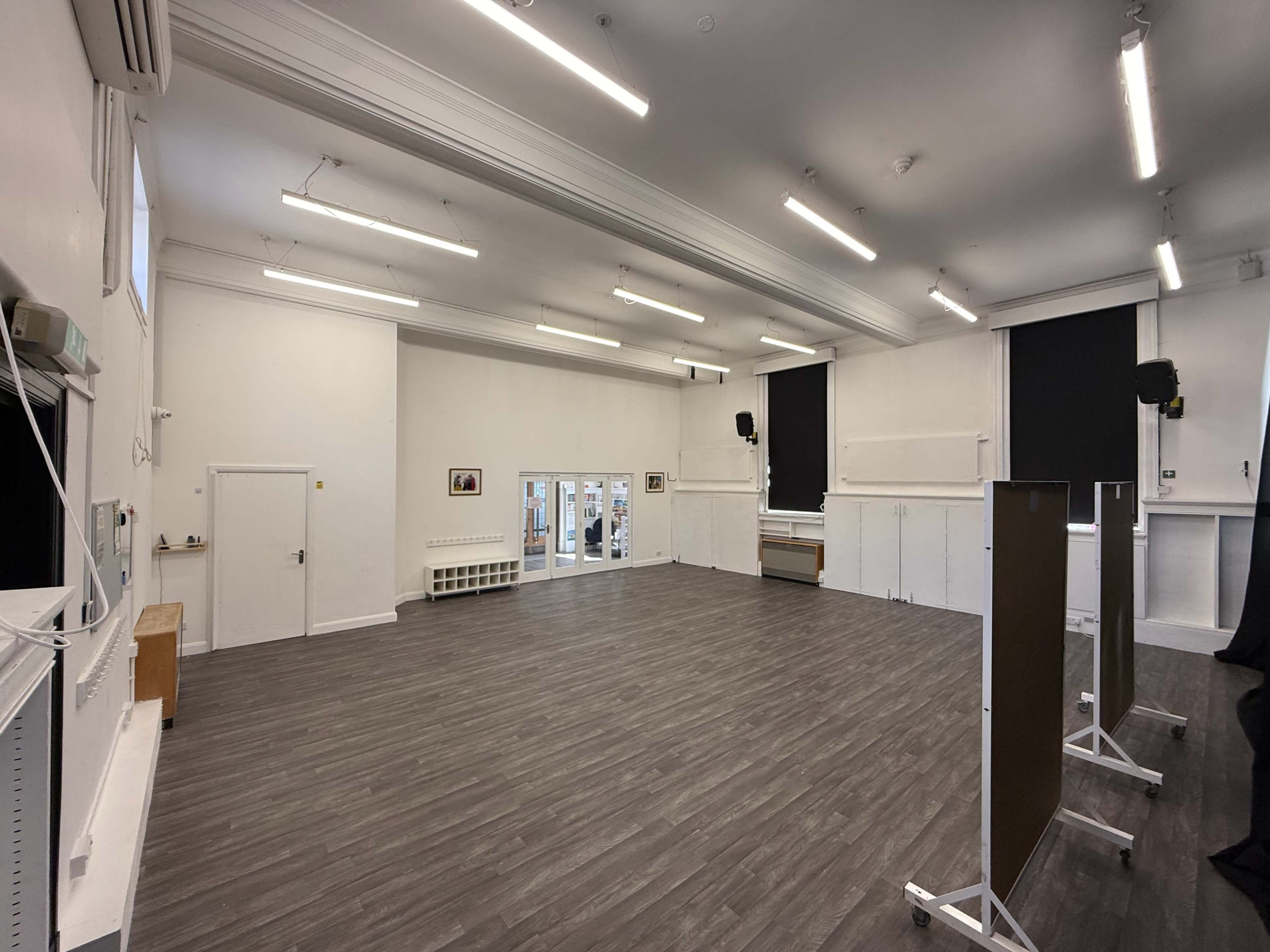 The image shows a large, empty room with wooden flooring, high ceilings, and white walls, featuring windows and mounted light fixtures.