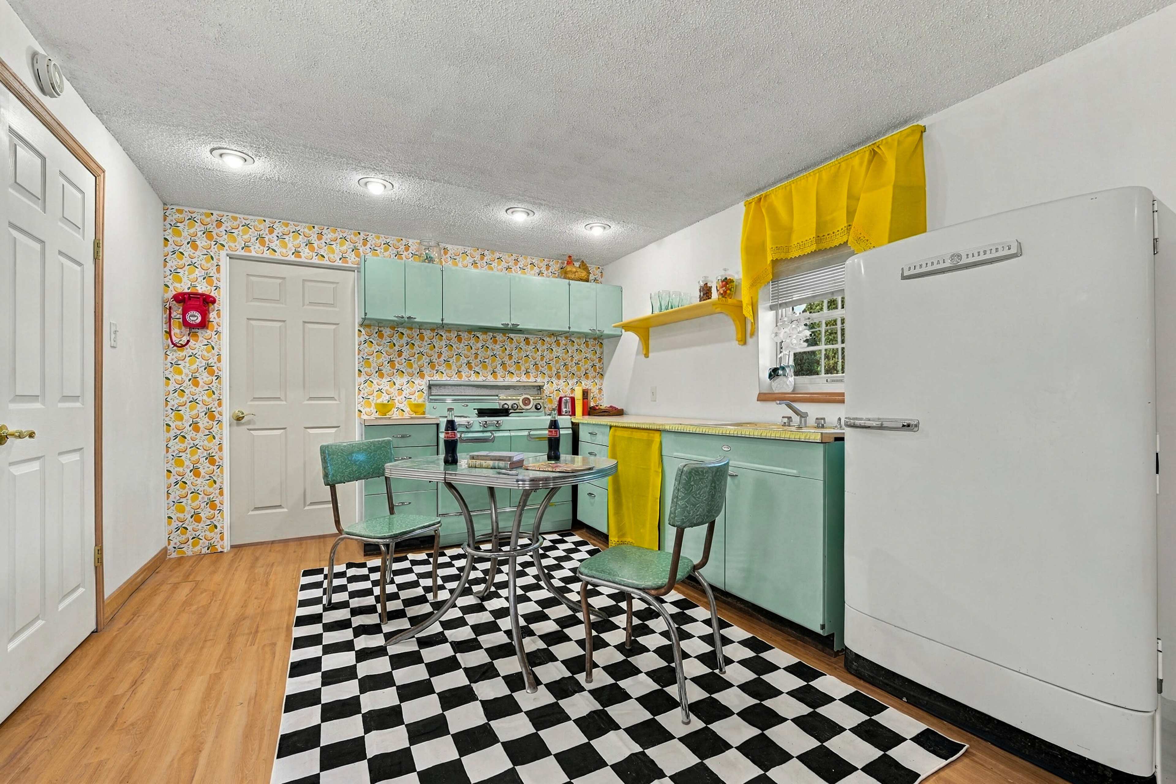 A vintage kitchen features mint green cabinets, a white retro refrigerator, a black and white checkered floor, and a small dining table with chrome and green chairs, accented by yellow curtains and floral wallpaper.