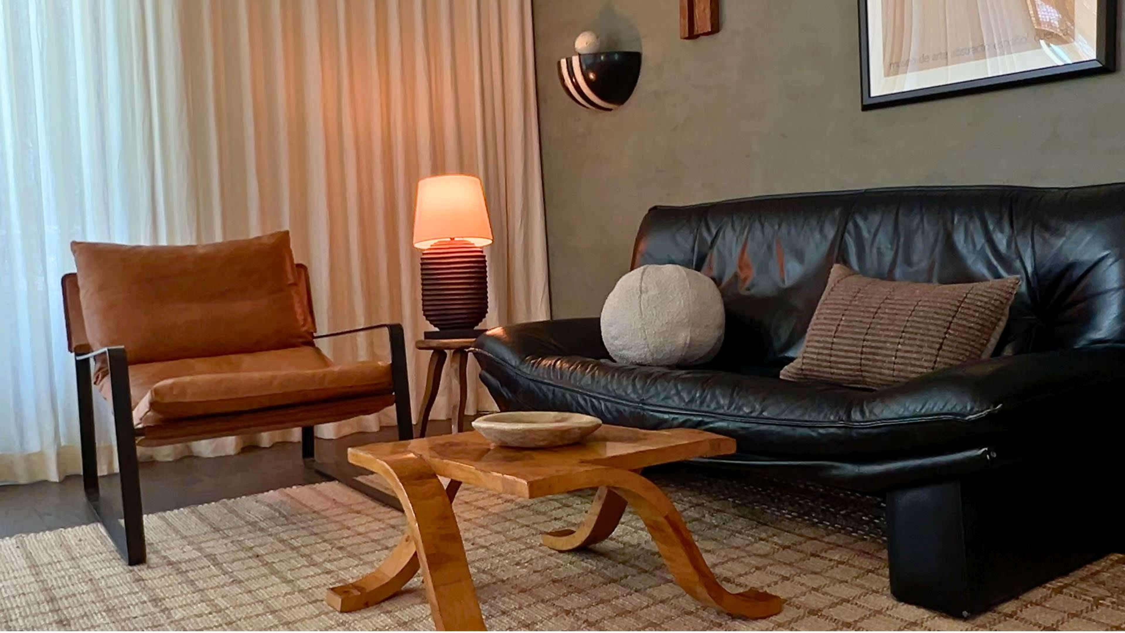The scene features a modern living room with a black leather sofa, a brown armchair, a wooden coffee table, and soft lighting from various sources.