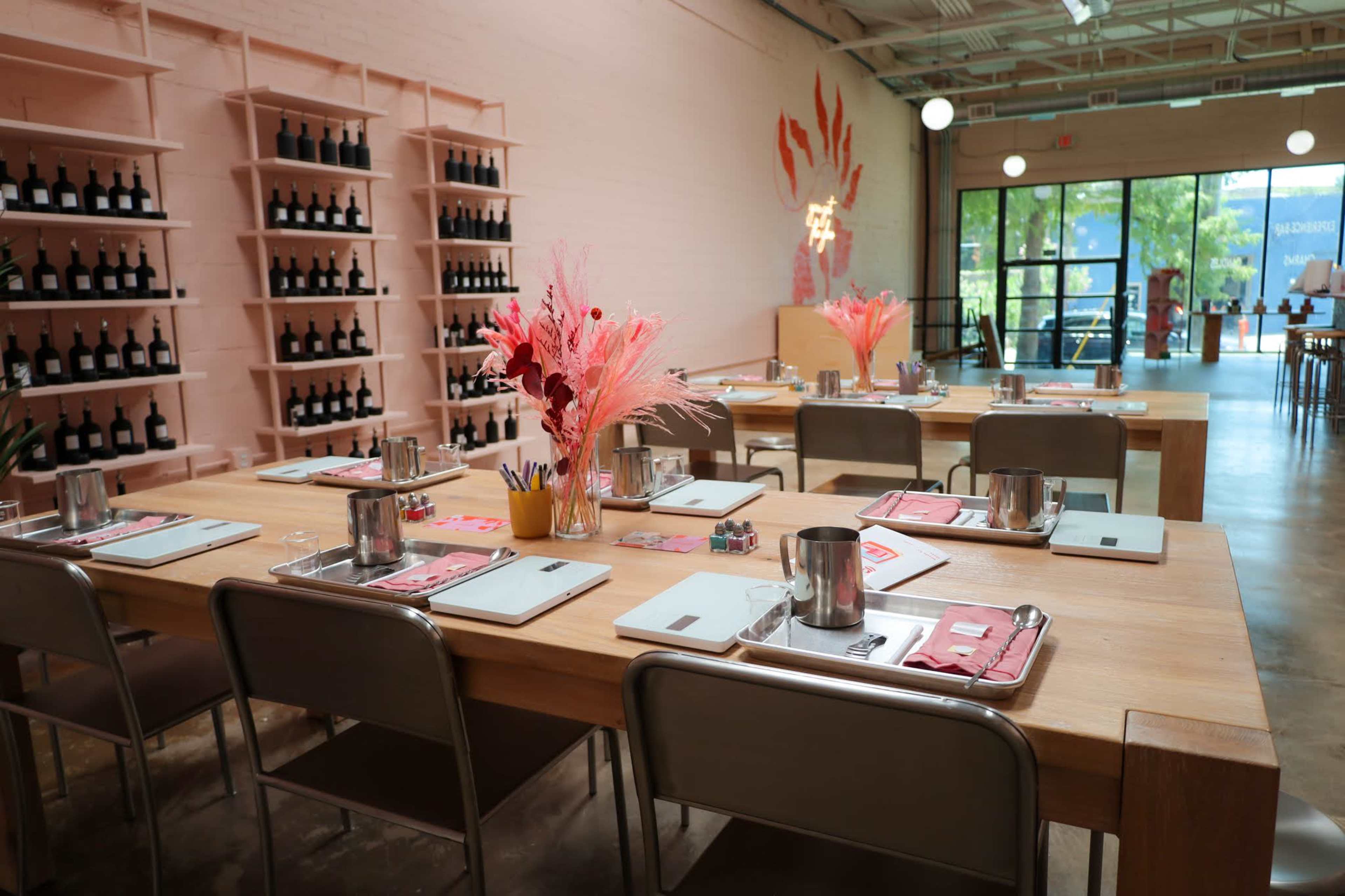 A dining area with wooden tables set for a meal, featuring wine bottles on shelves and decorated with pink floral arrangements.