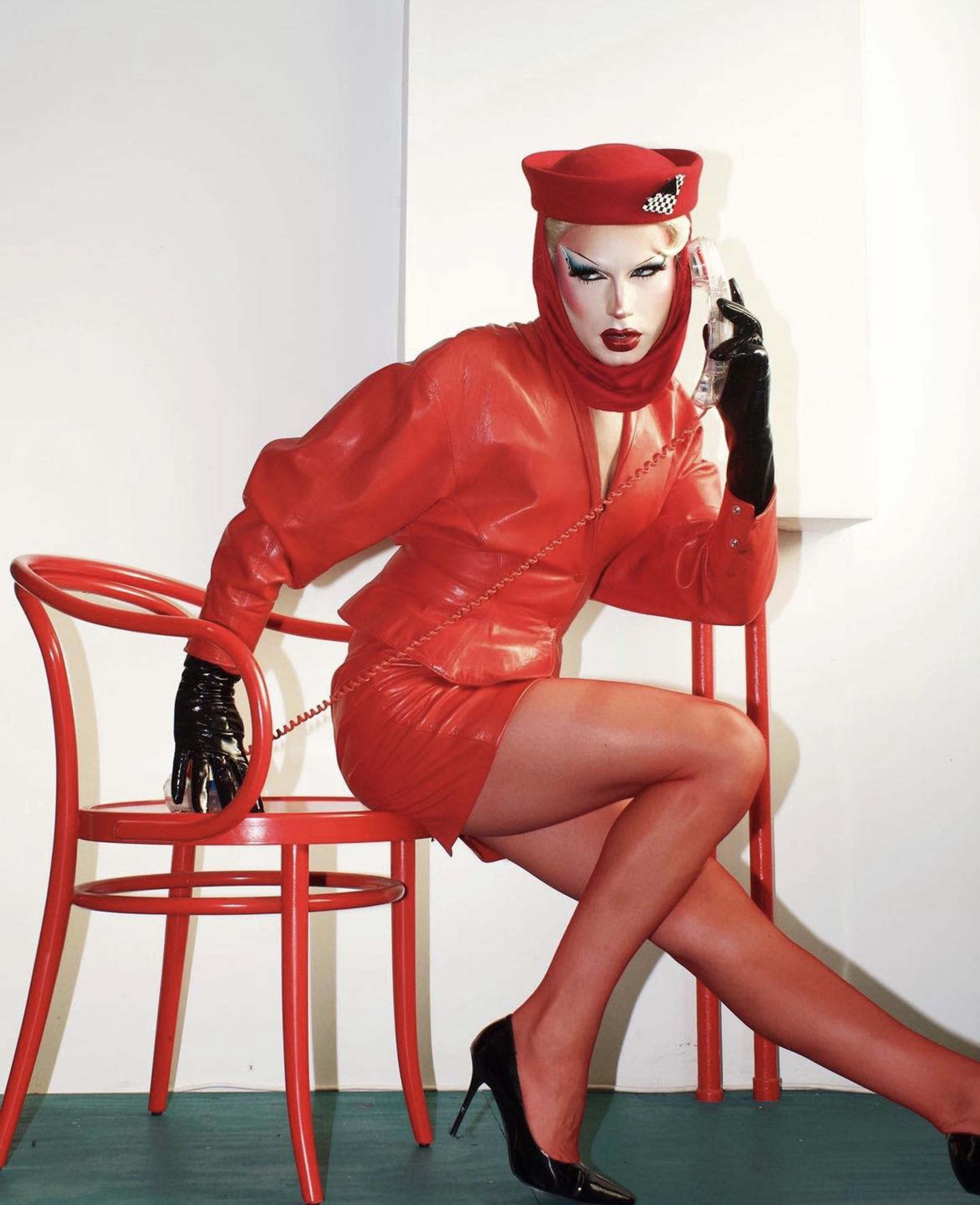 A figure in a red latex outfit sits on a red chair while holding a vintage telephone.
