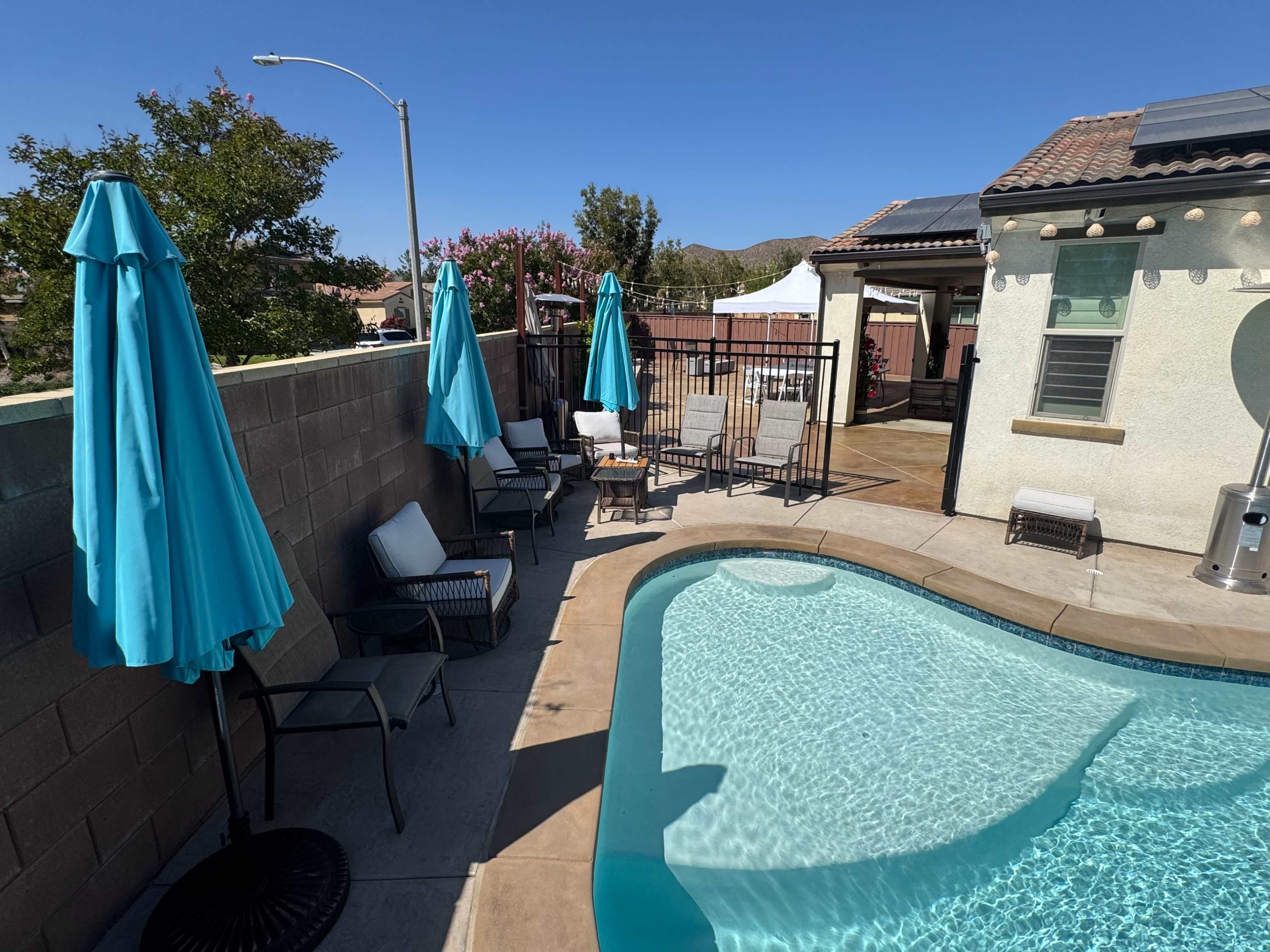 The image shows a backyard with a swimming pool surrounded by lounge chairs and umbrellas on a sunny day.