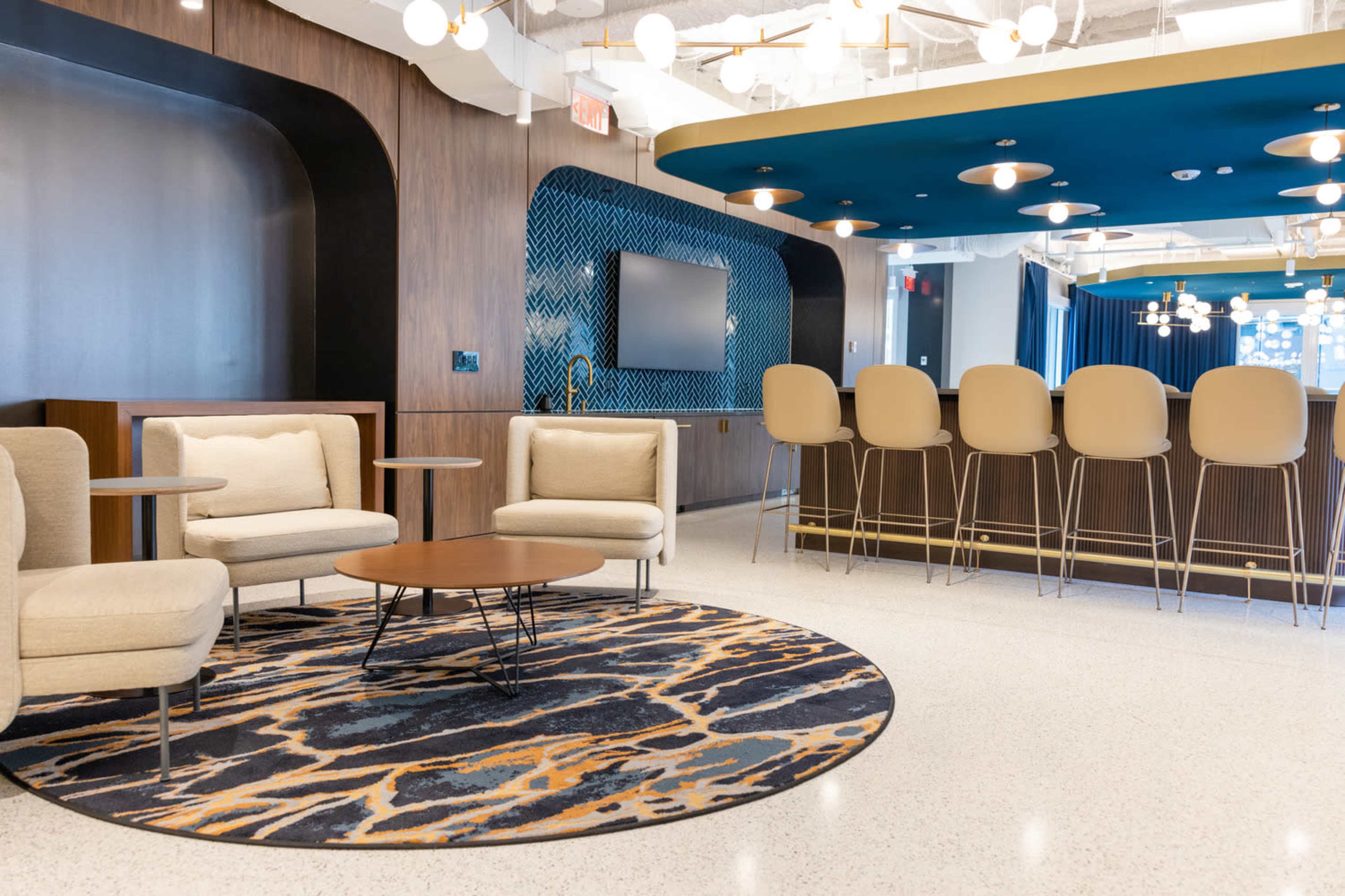 A modern lounge area with beige upholstered seating around a round coffee table, a patterned rug, and a bar counter with high stools in a bright, airy space.