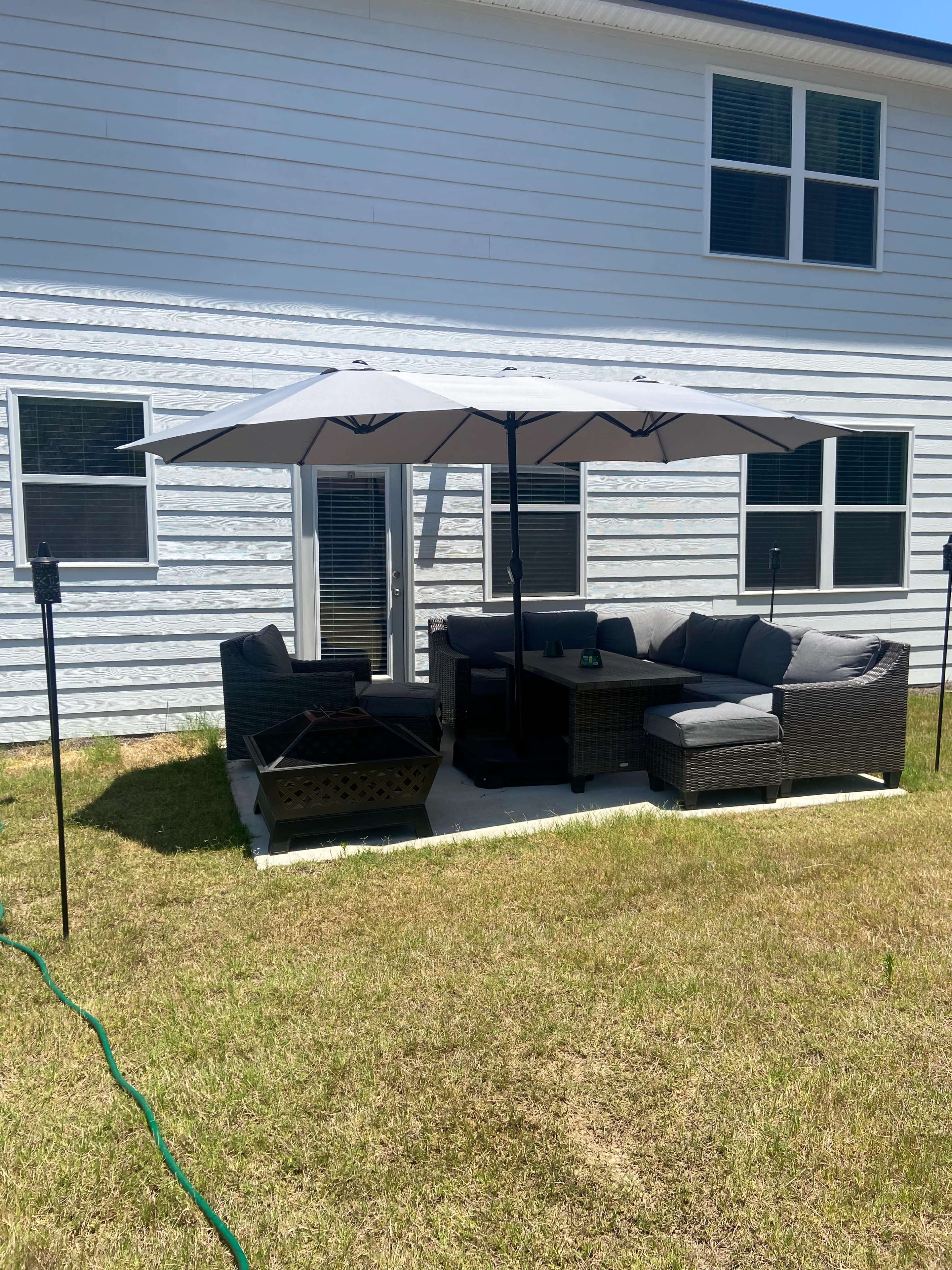 A patio area featuring a sectional sofa and an umbrella is located in a grassy backyard next to a house.