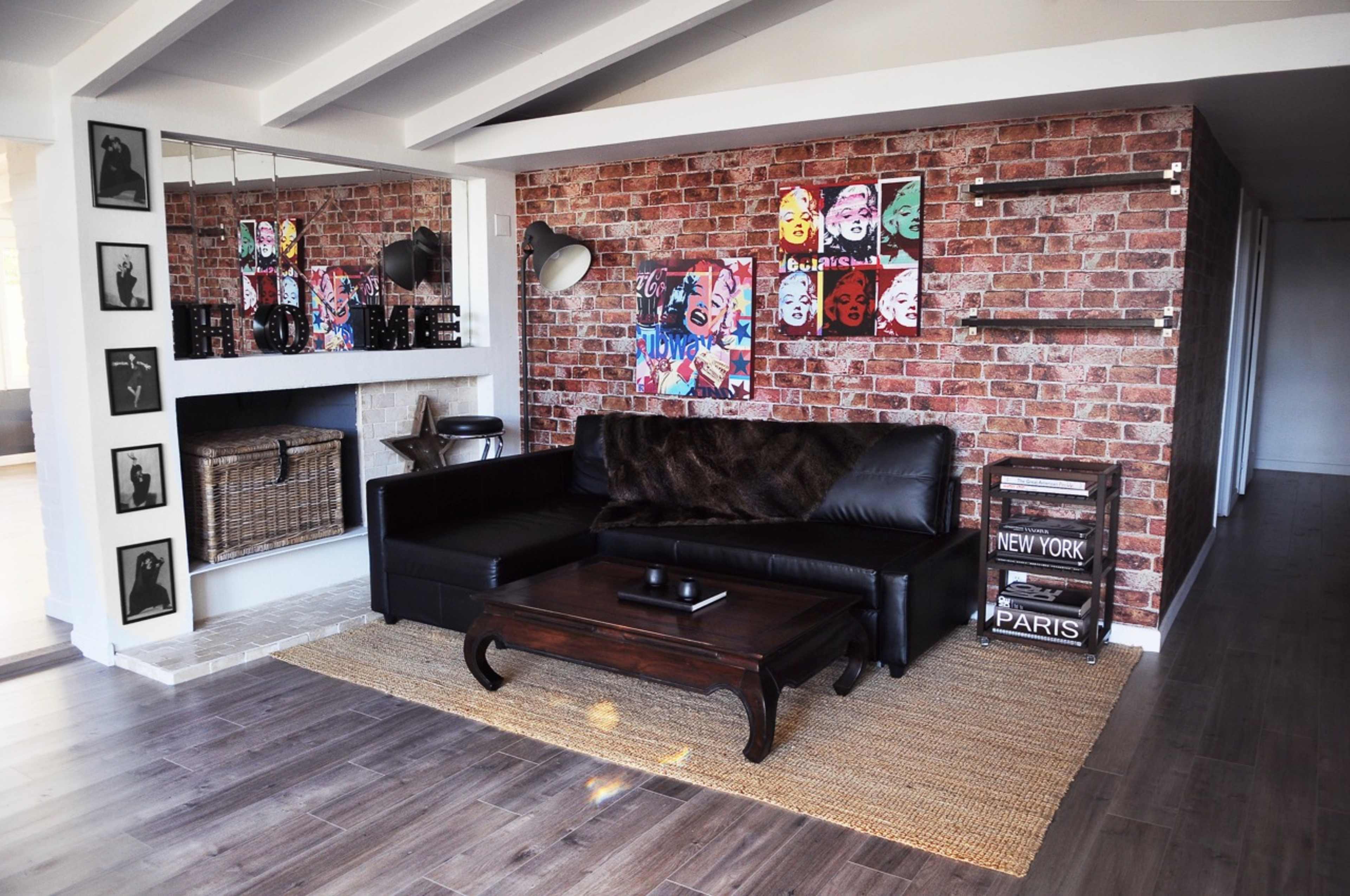 The image shows a modern living room with a dark leather sofa, a wooden coffee table, and a textured brick wall adorned with colorful artwork and framed photographs.