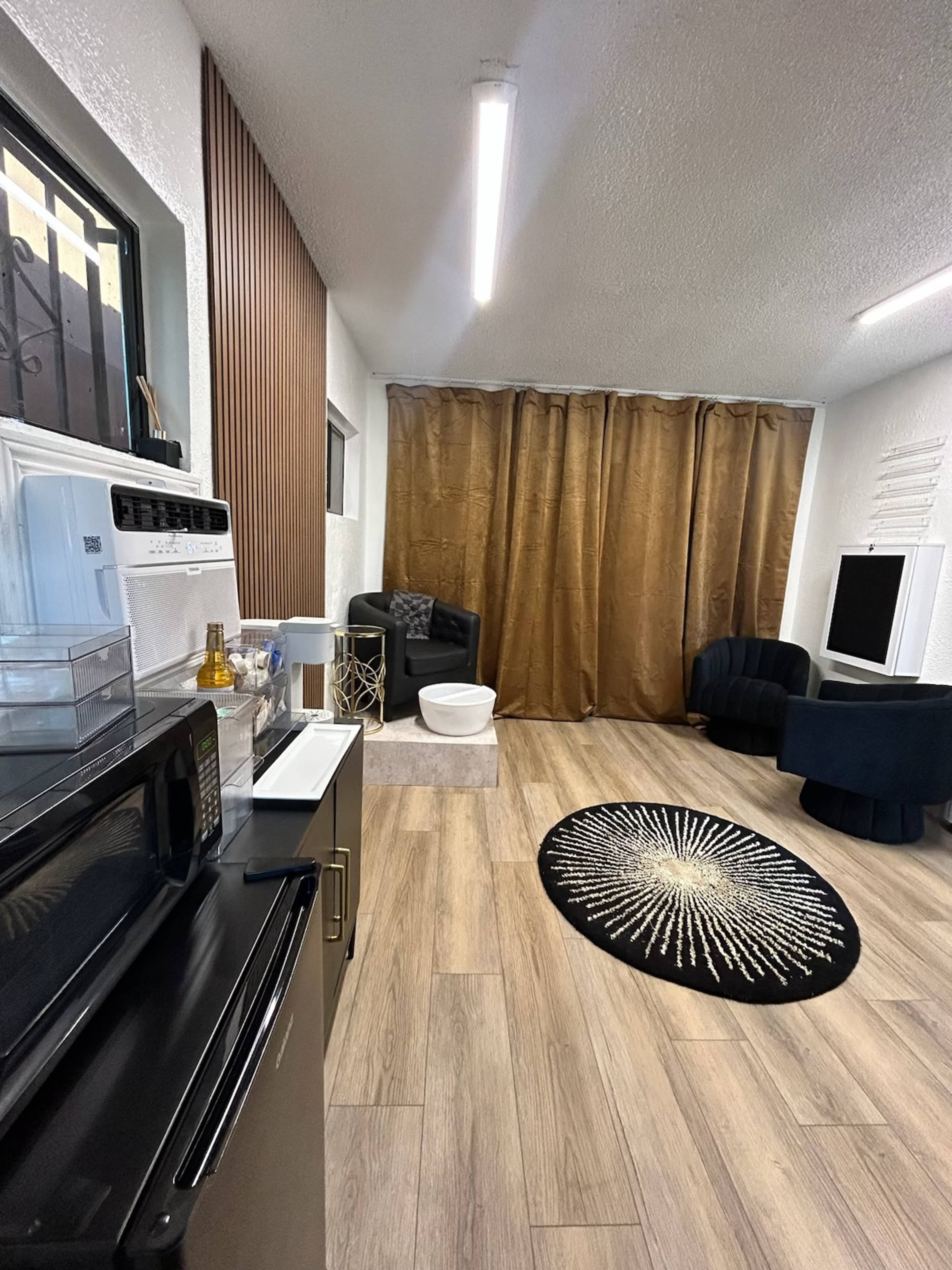 The image shows a modern room featuring a kitchenette with a microwave and a decorative rug, alongside a seating area with a black armchair and a white stool, separated by a brown curtain.