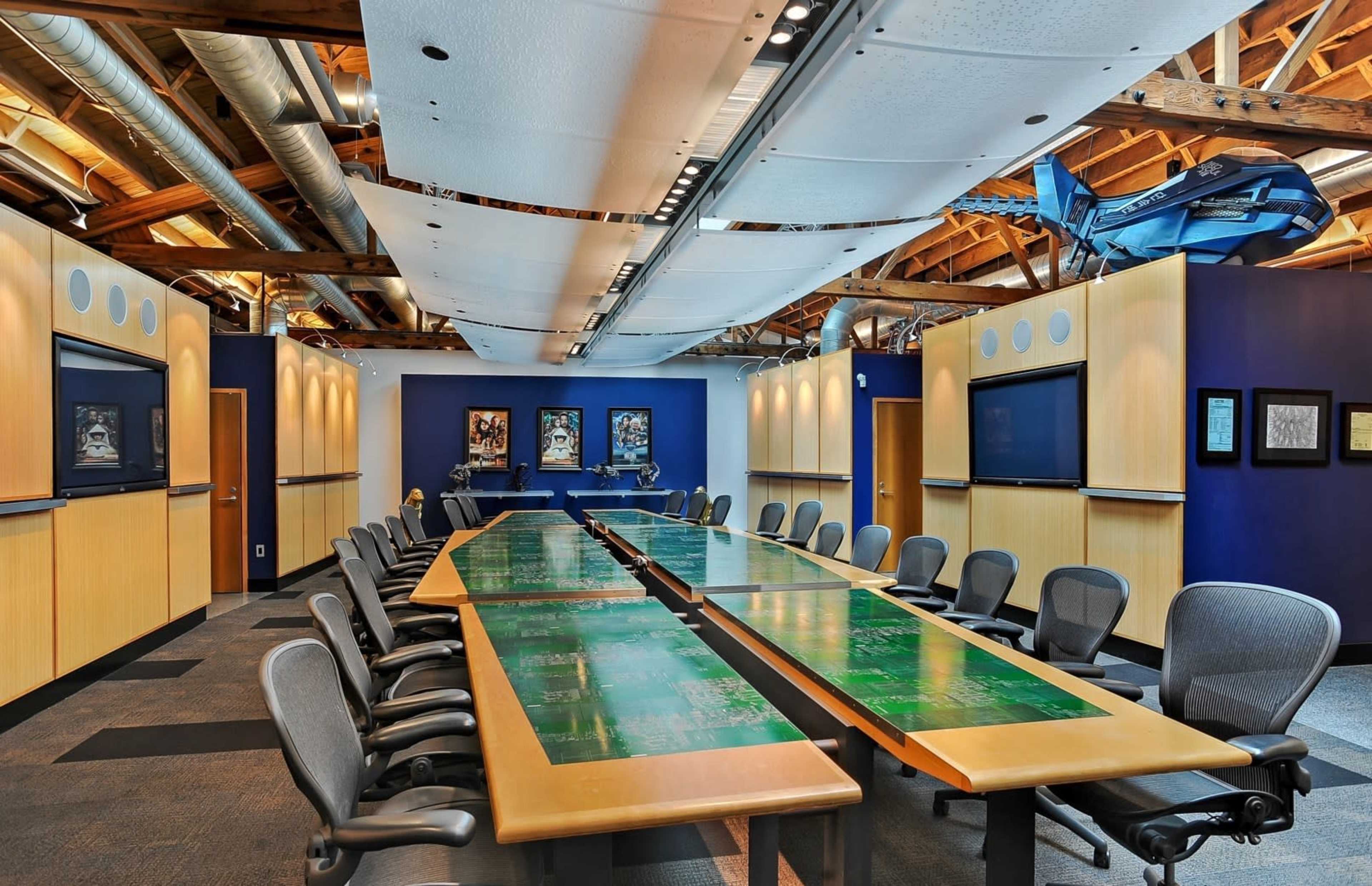 A large conference room features a long table with green graphic designs, surrounded by ergonomic chairs, and is equipped with screens on the walls.