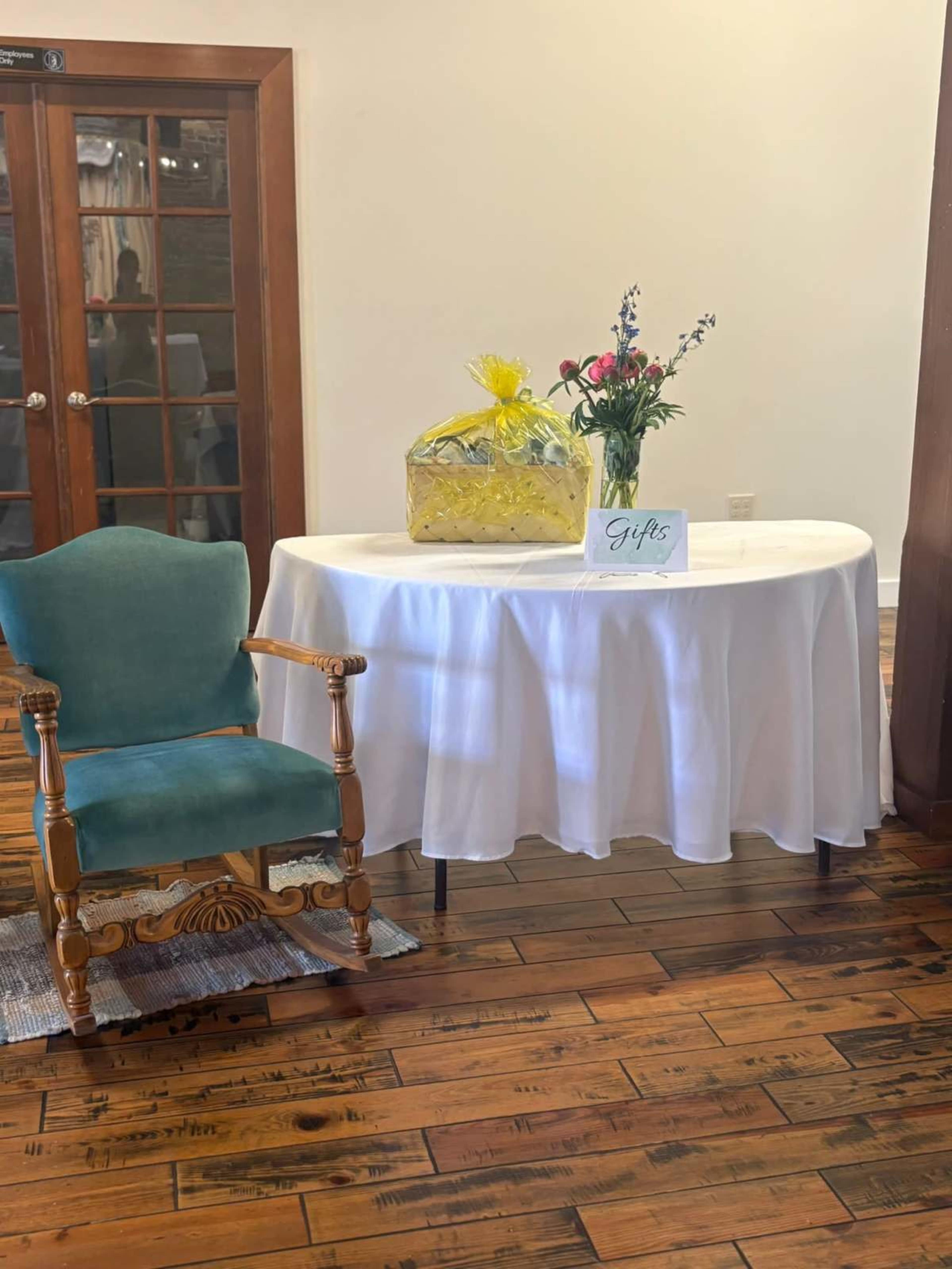 A small table covered with a white tablecloth displays a gift wrapped in yellow cellophane alongside a bouquet of flowers and a "Gifts" sign, set in a room with wooden flooring and a chair nearby.