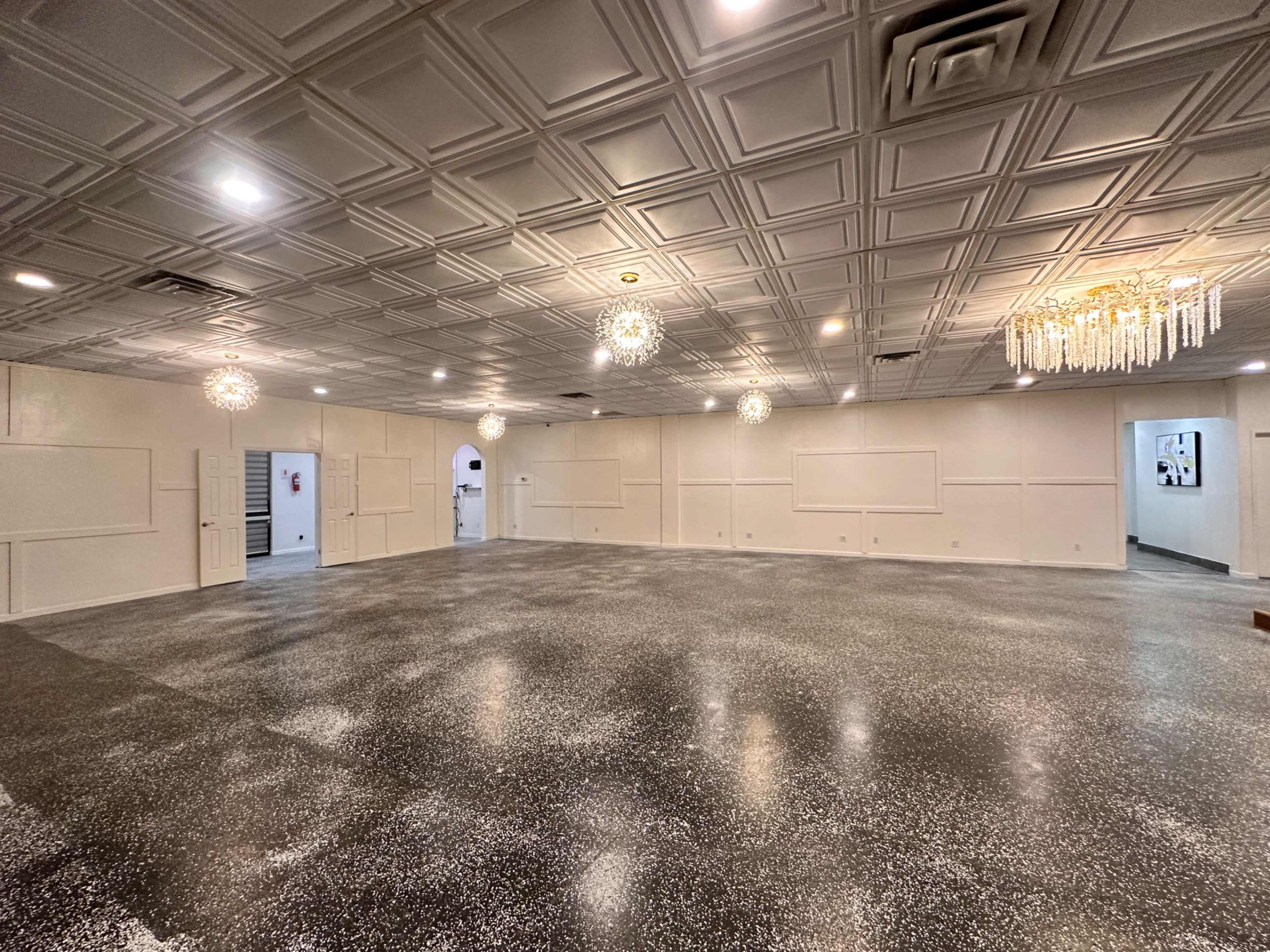 A spacious, empty room with a decorative ceiling and two distinct styles of chandeliers illuminating the floor covered in a speckled finish.