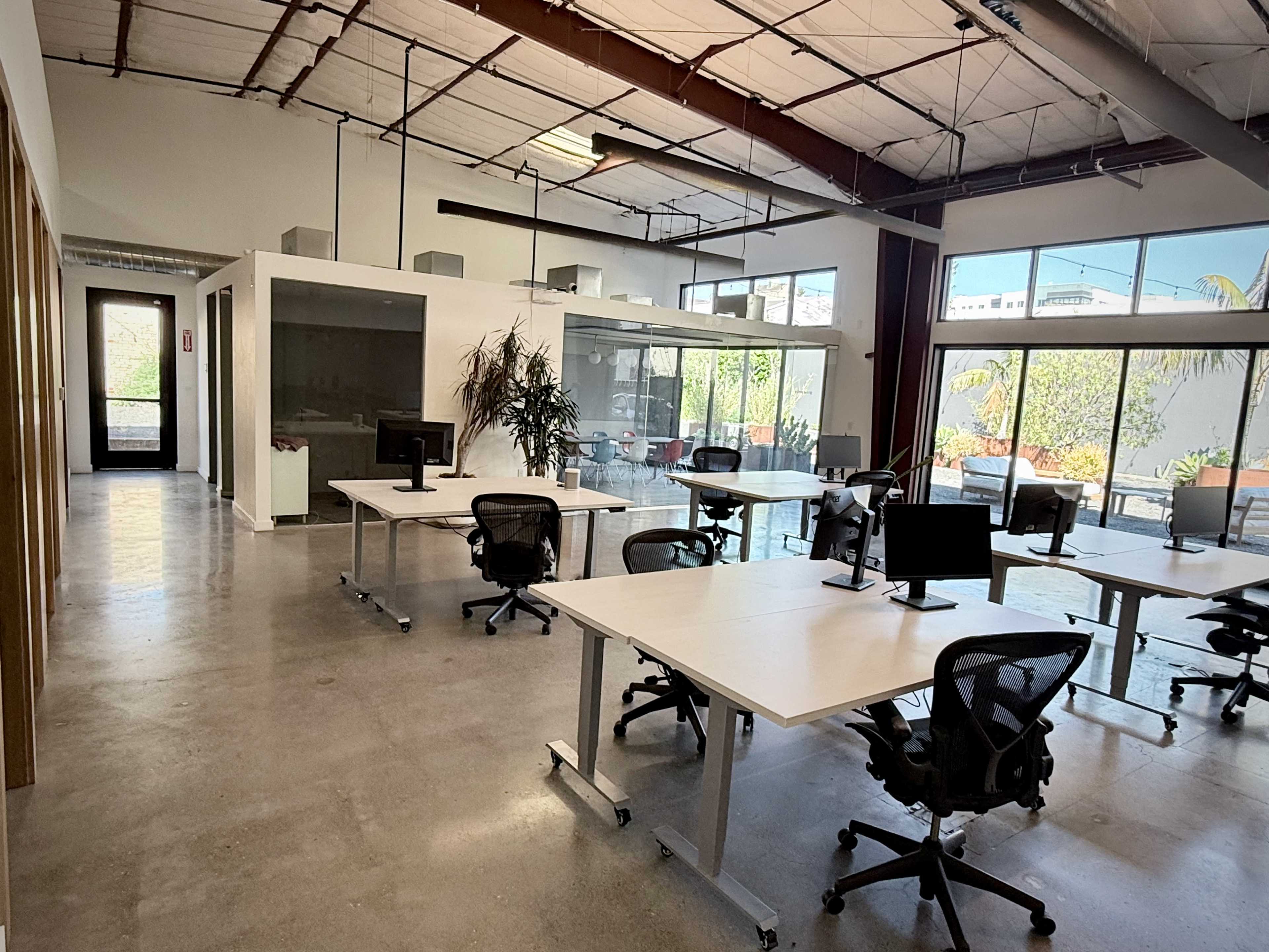 The image shows a modern, open-plan office with several workstations equipped with computers and large windows allowing natural light to enter.