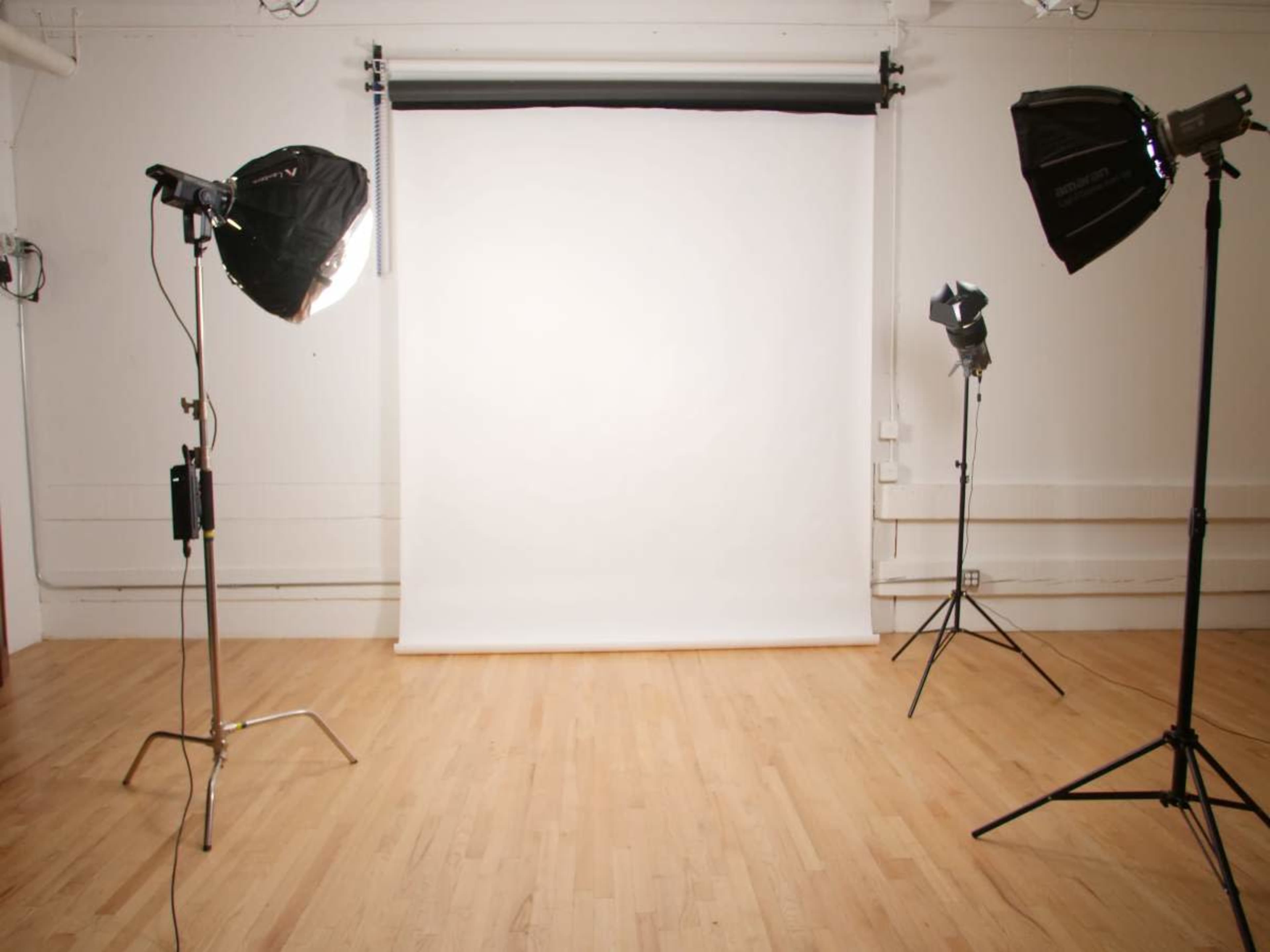 The image shows a photography studio with a blank white backdrop and three lighting setups on stands.