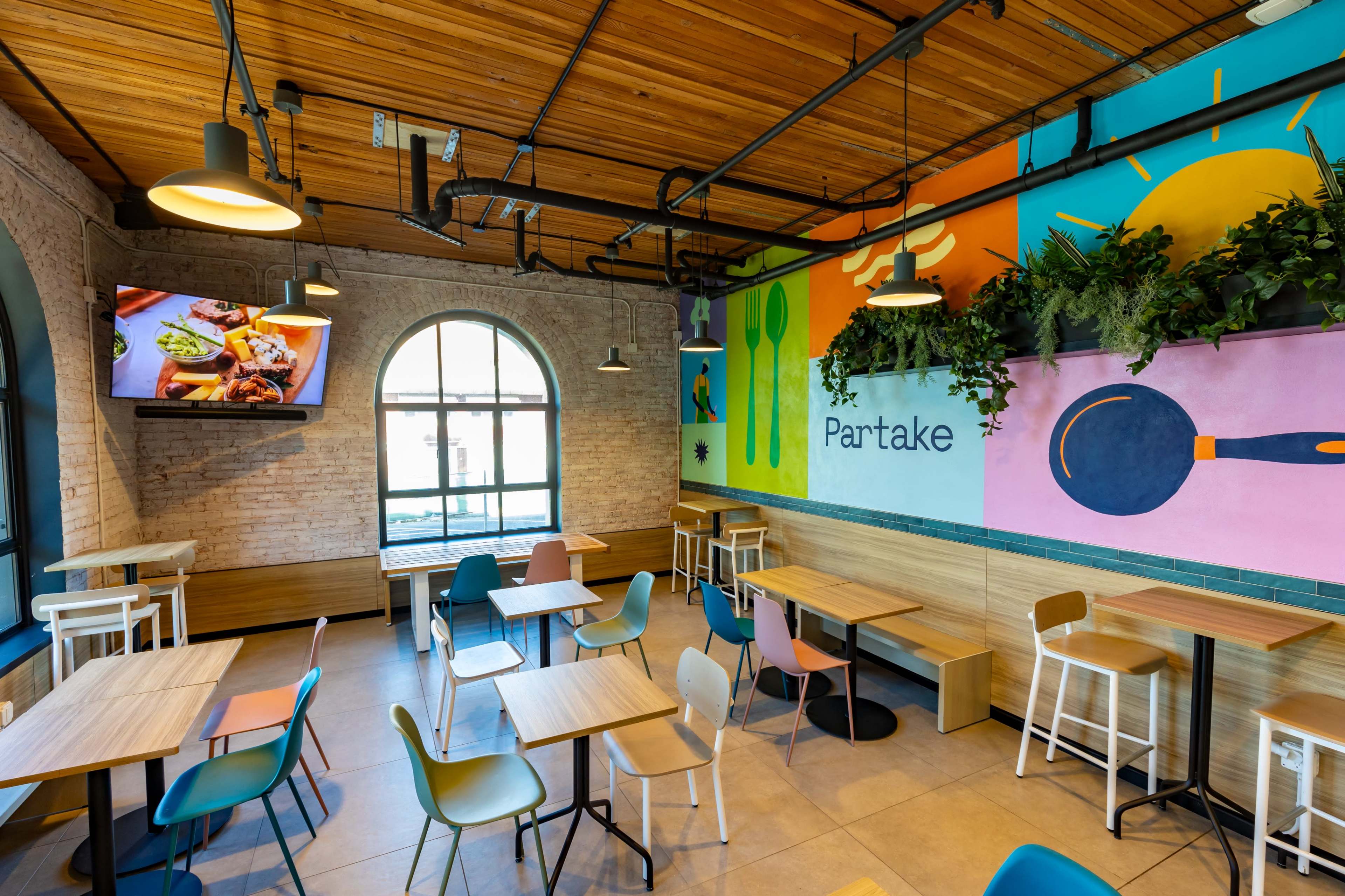 The image shows a modern restaurant interior with wooden ceilings, colorful wall art, and a mix of seating options including tables and chairs.