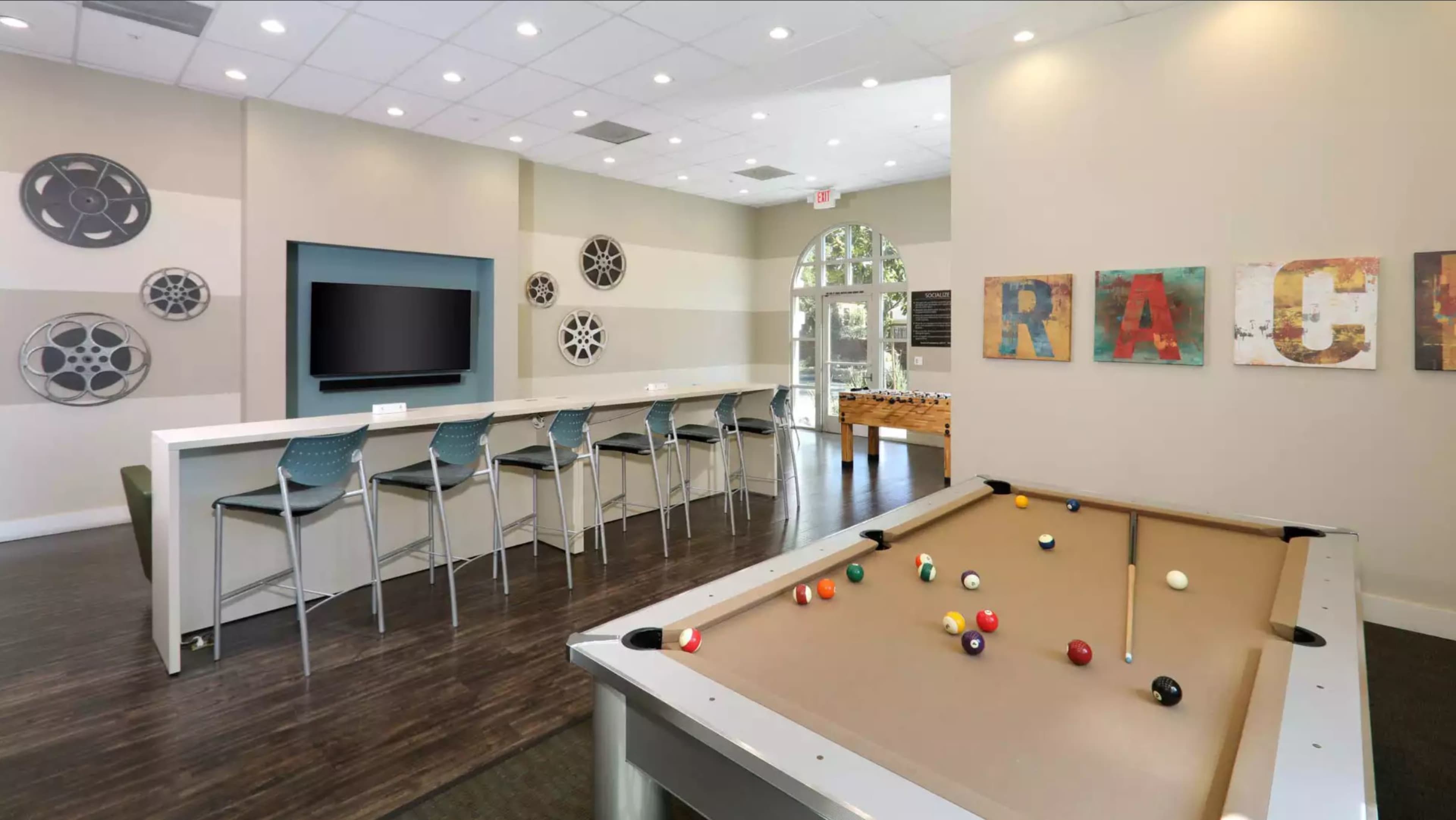 A recreational room with a pool table in the foreground, a bar area with high stools, and a large TV on the wall, decorated with art pieces spelling "RACE."