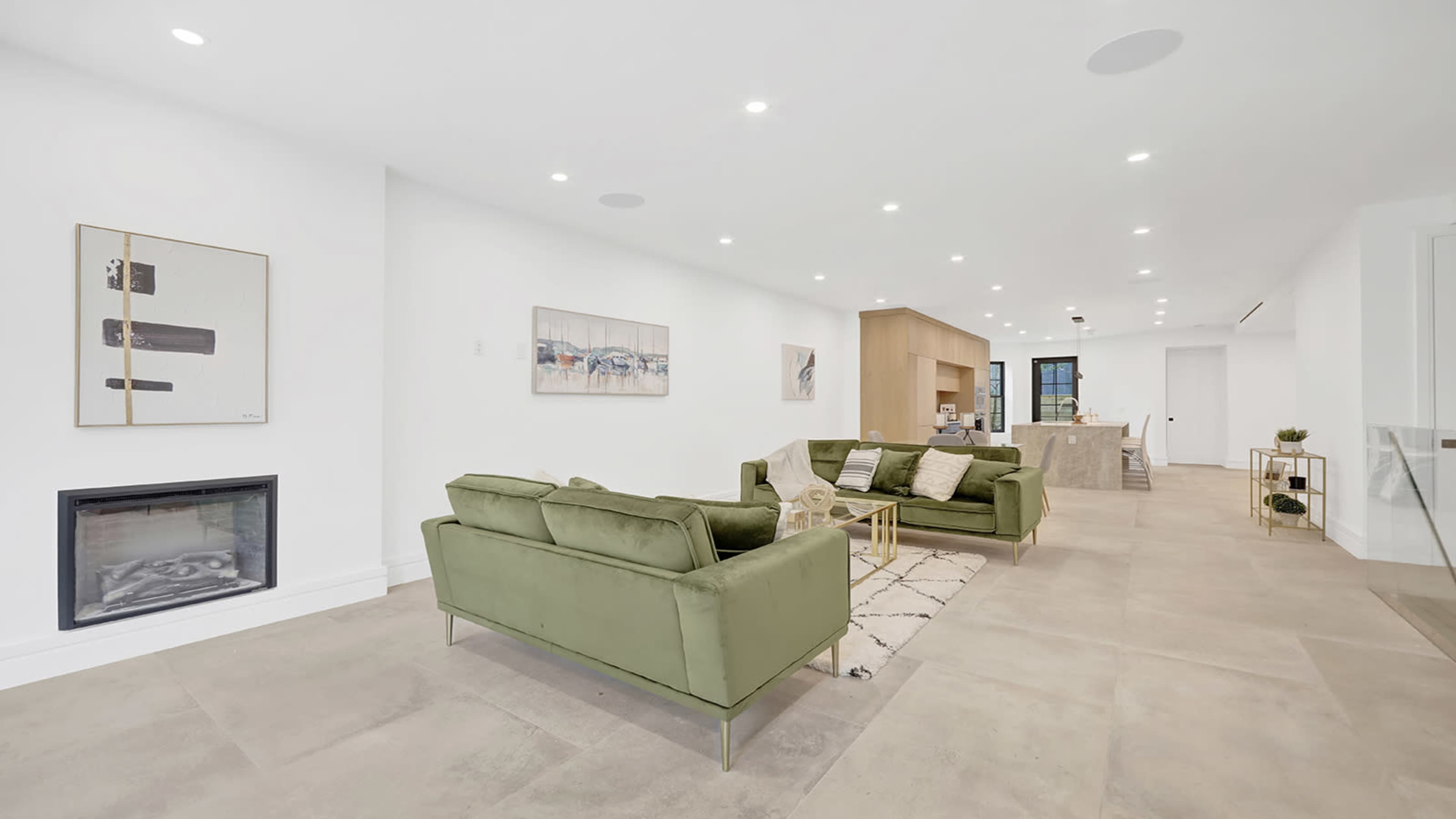 A spacious modern living room features green sofas, a minimalist artwork on the wall, and an open layout connecting to a kitchen area.