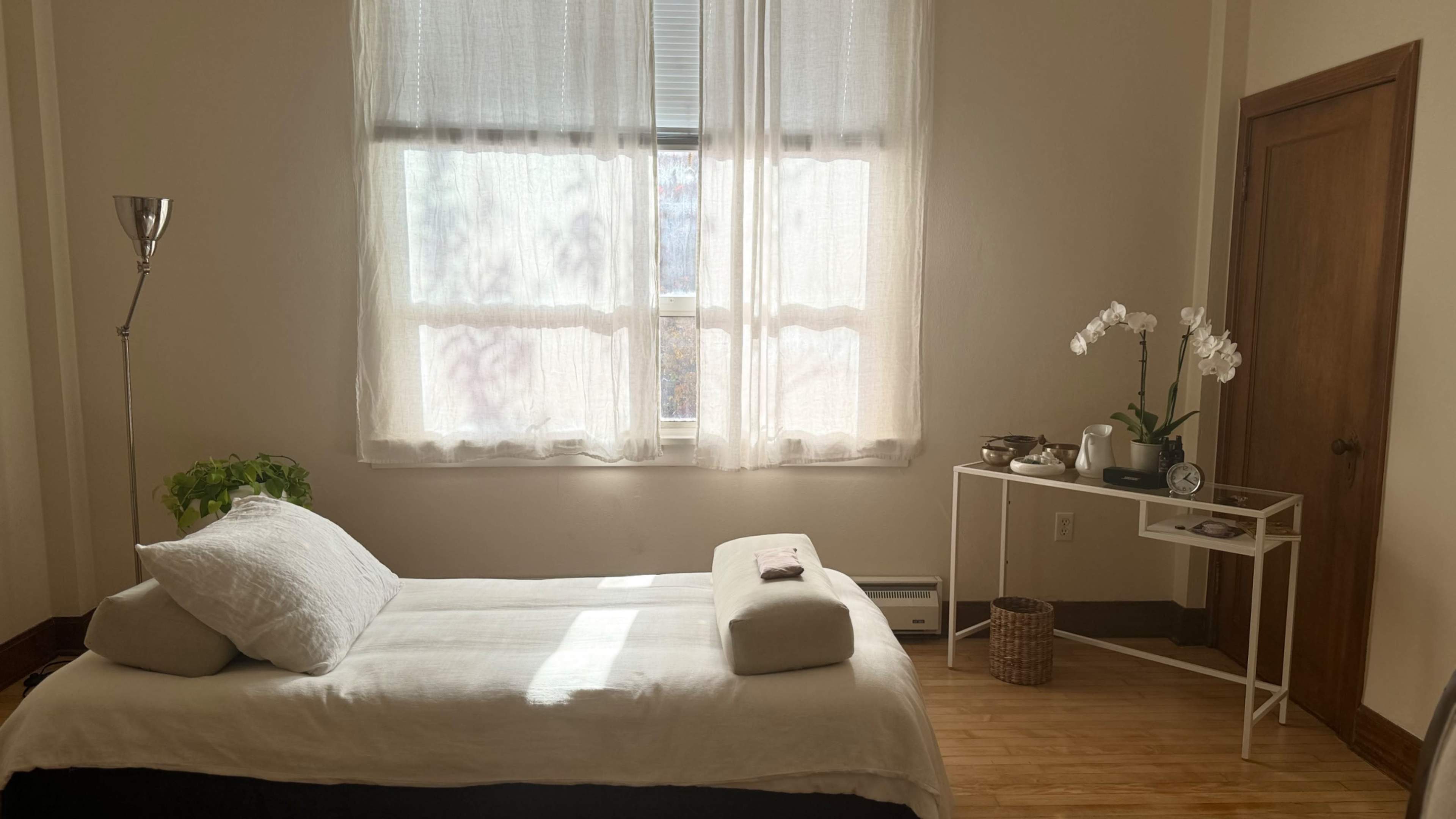 The image shows a bright, minimalistic bedroom with a bed, a small table, and a decorative plant near a window with sheer curtains.