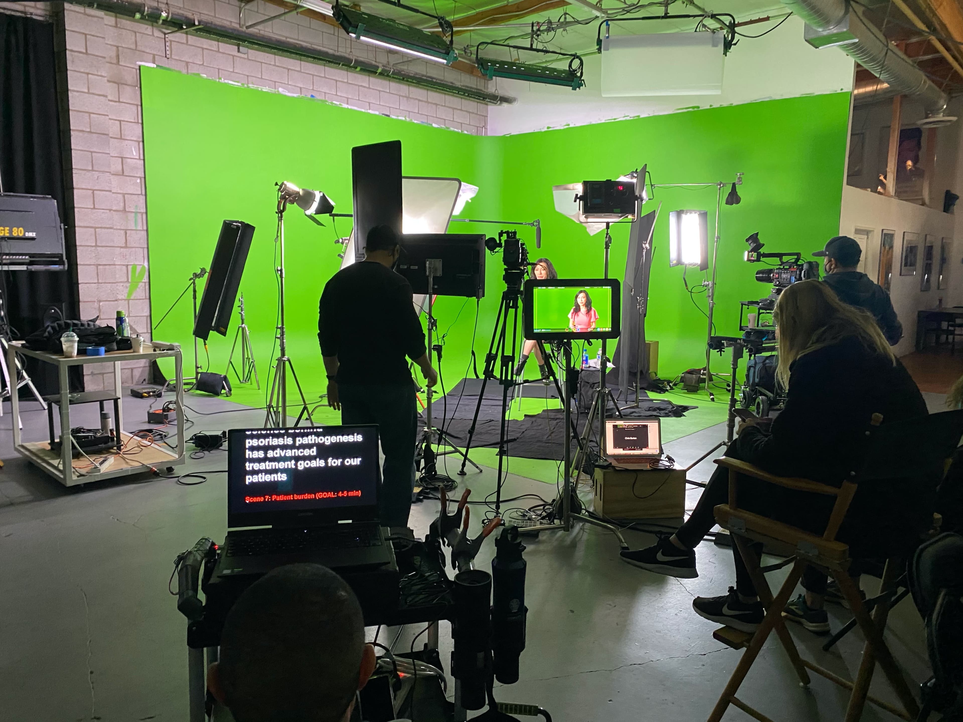A video production setup with multiple cameras, lights, and a green screen, featuring a speaker in front of a monitor and crew members preparing to film.