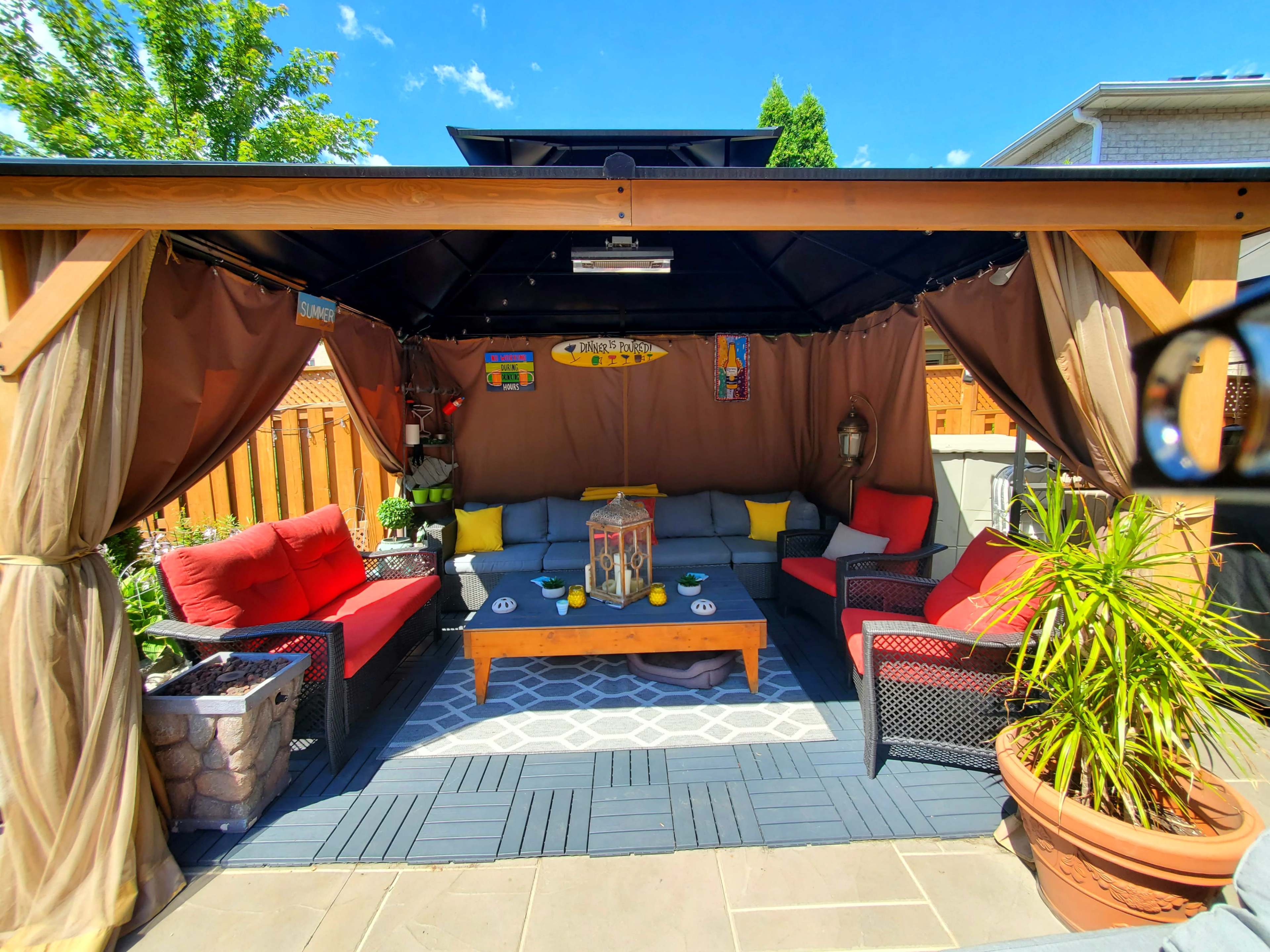 A covered outdoor seating area features red and yellow cushions on black wicker furniture, a wooden coffee table, and potted plants under a tent-like structure.