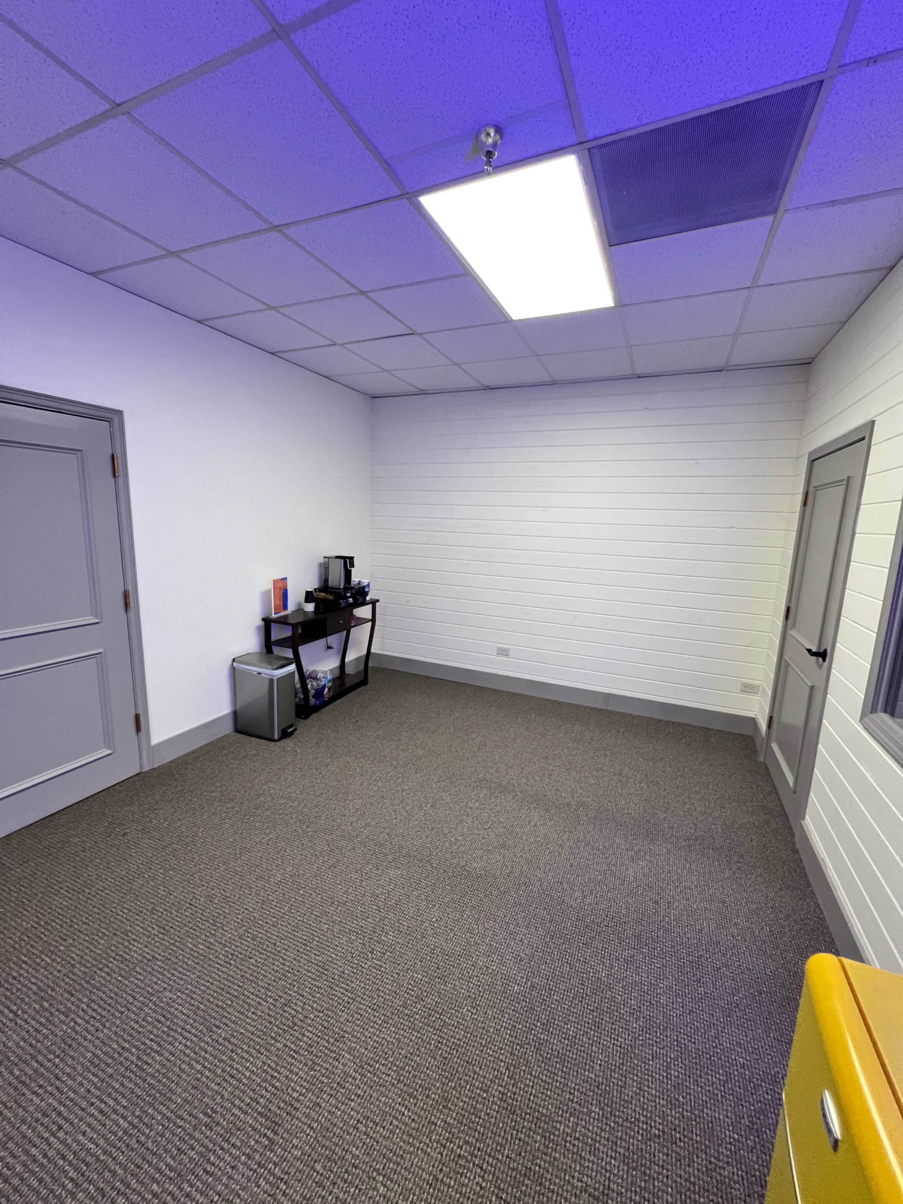 The image shows an empty office space with light-colored walls, a carpeted floor, and minimal furnishings including a small table with a coffee maker and a filing cabinet.