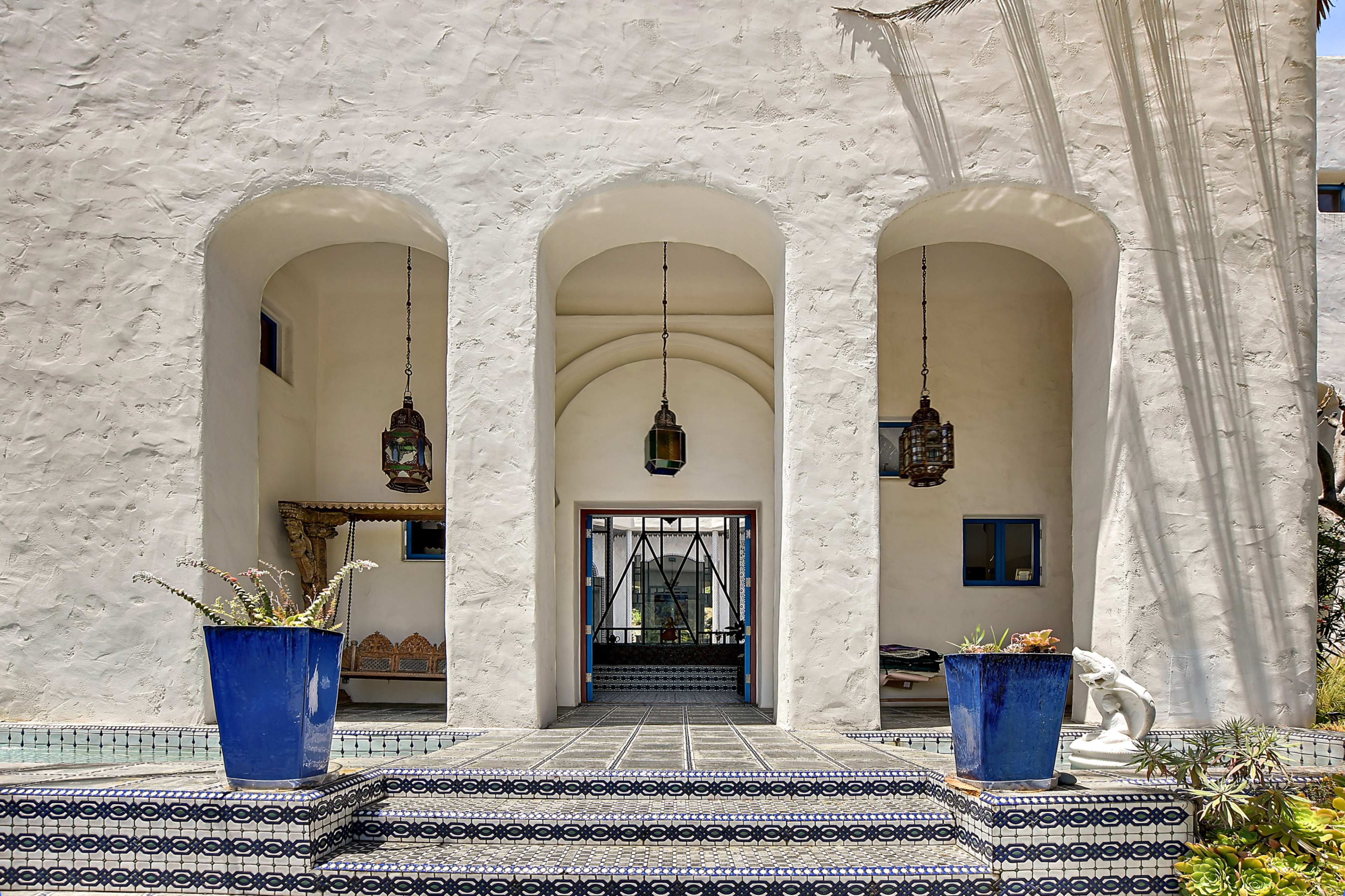 The image shows a large, white stucco entrance with three arched openings, flanked by two blue planters and featuring hanging lanterns.